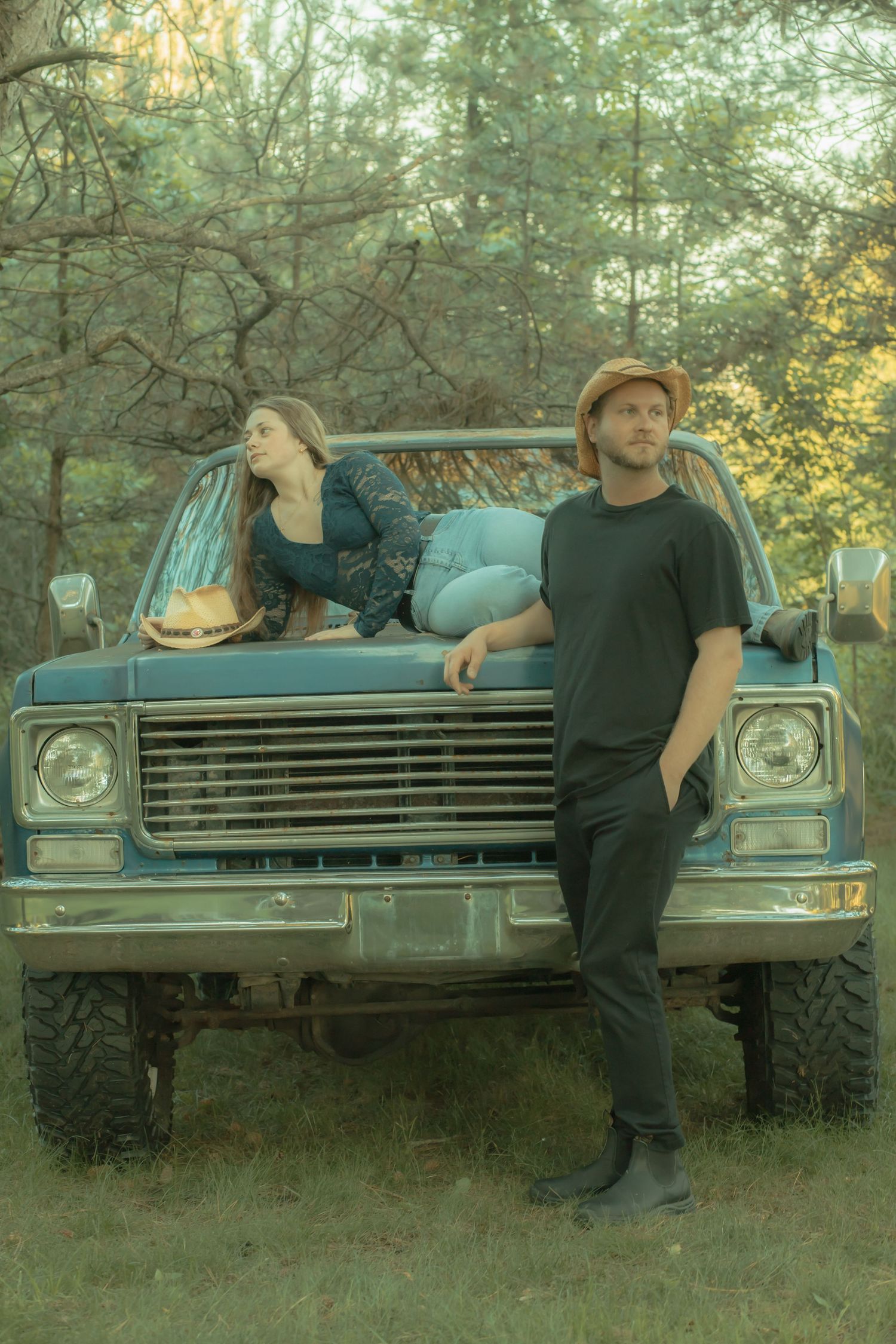Man and woman posing with vintage blue pickup truck in a forest setting.