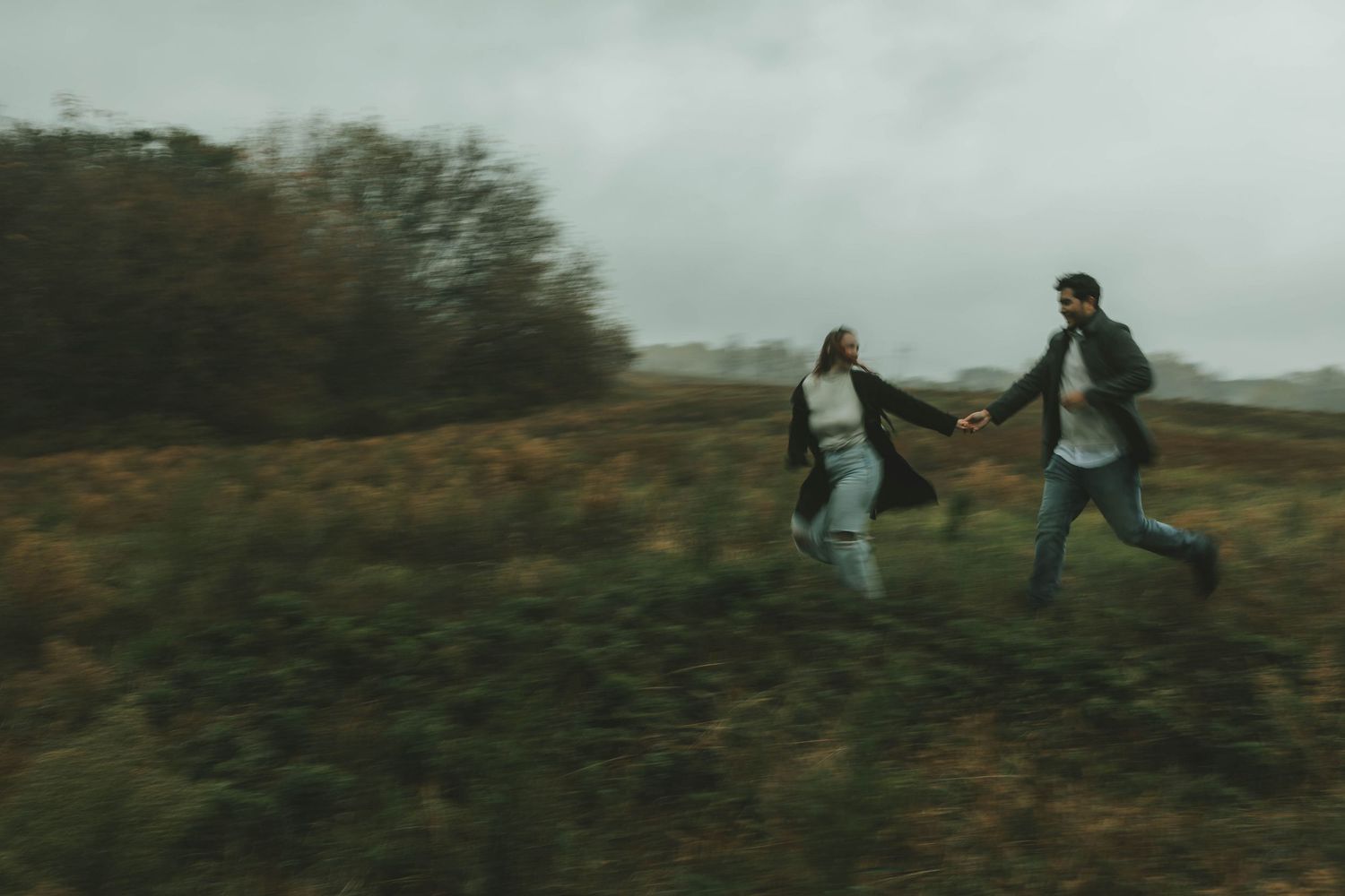 A couple playfully runs through a misty field together on an overcast autumn day.