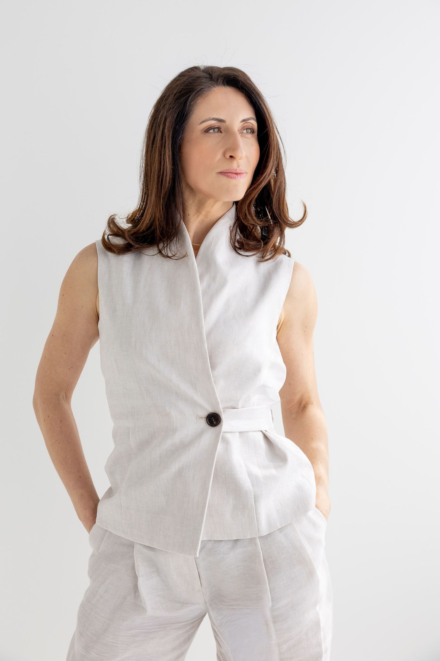 Woman in sleeveless white linen vest and matching trousers standing with hands in pockets against light background.