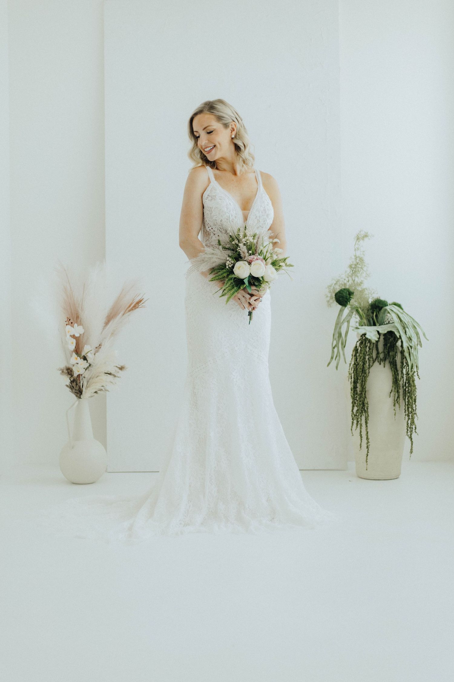 A bride in a fitted lace wedding gown holds a soft pastel bouquet against a white backdrop with pampas grass decor.