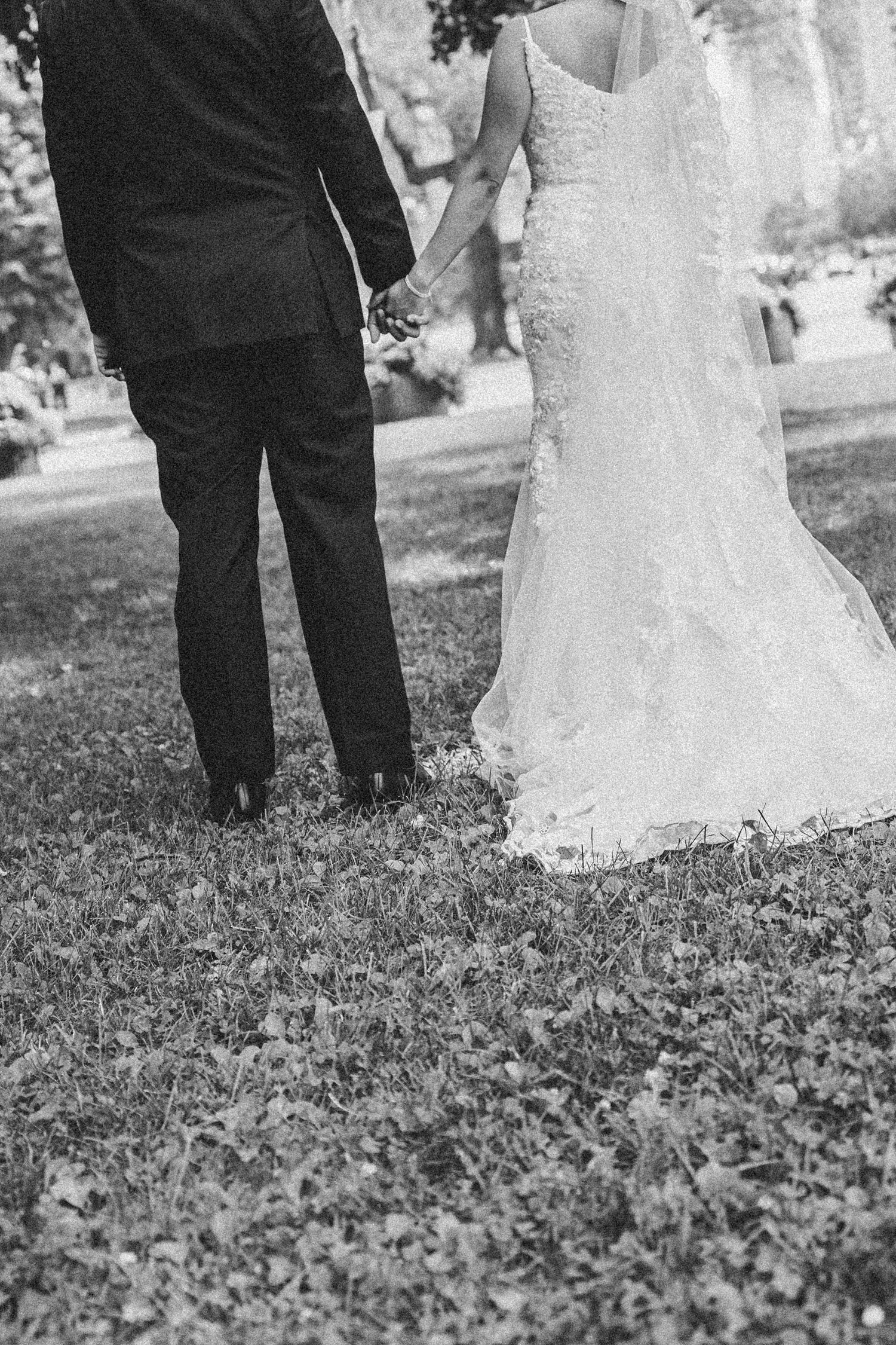 Black and white wedding photo showing groom in tuxedo and bride in lace dress standing together on grass.