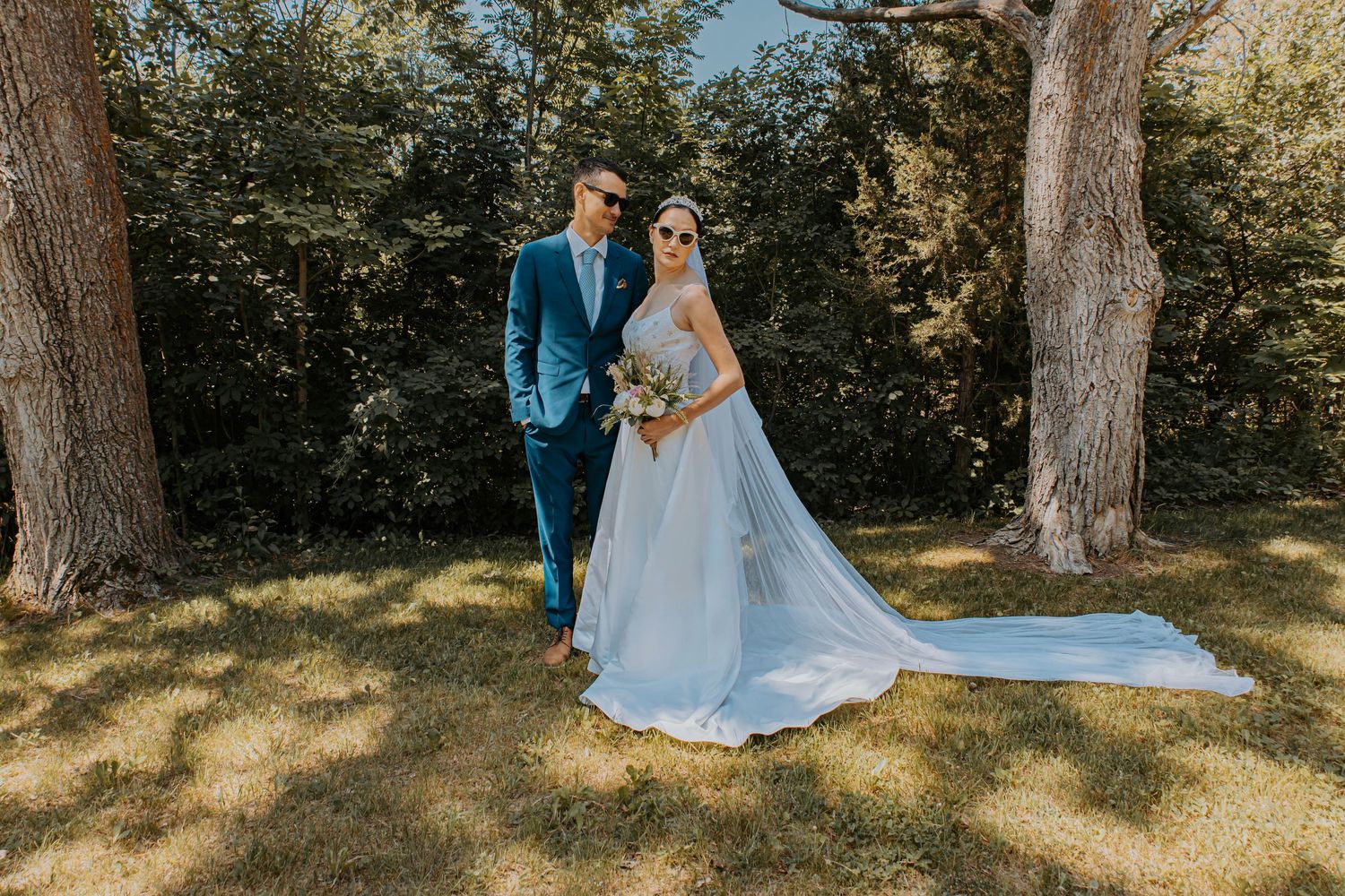 A newlywed couple stands together in a sunny garden setting with large trees and dappled sunlight.