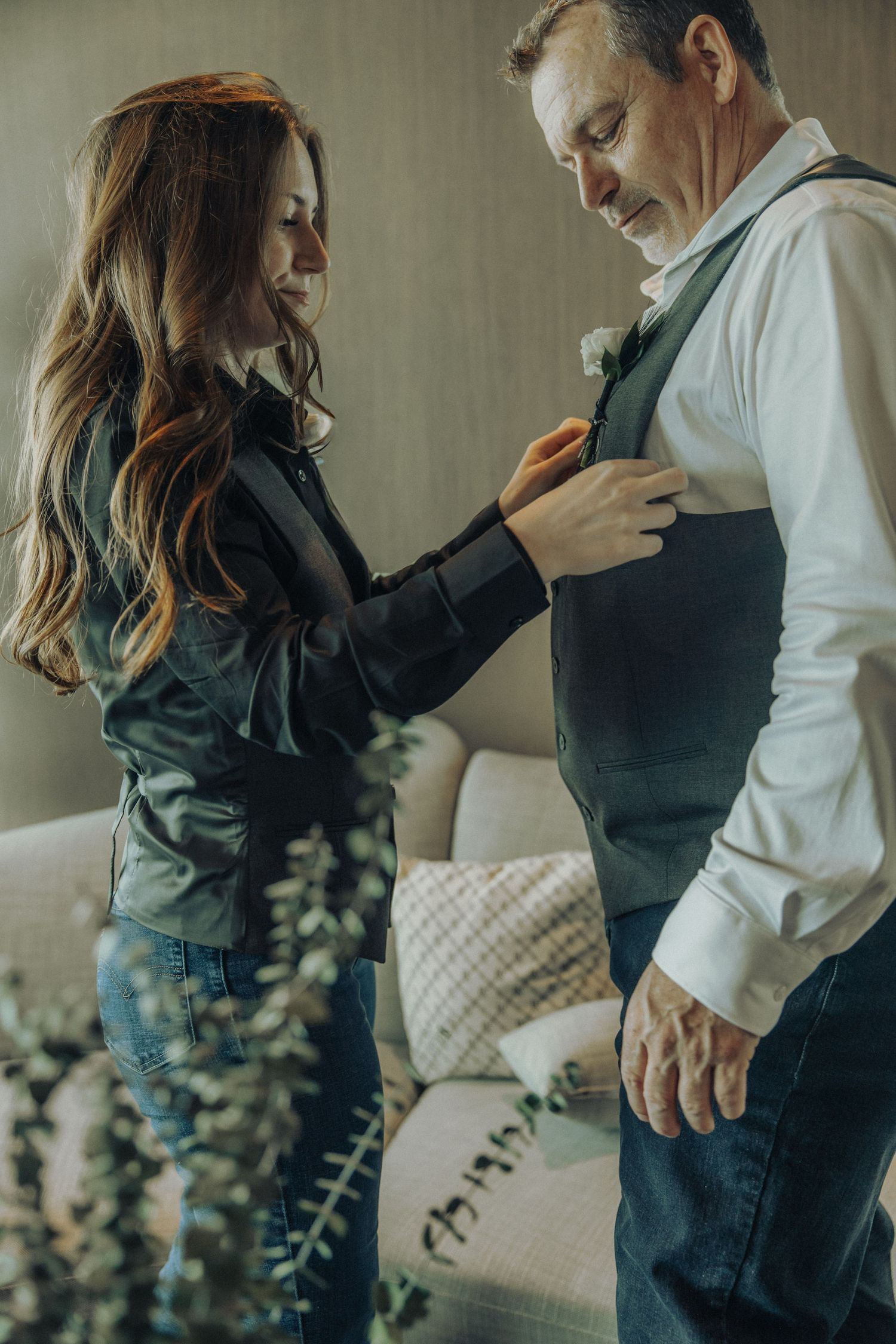 Bride helping groom adjust his vest boutonniere before wedding ceremony in a cozy indoor setting.