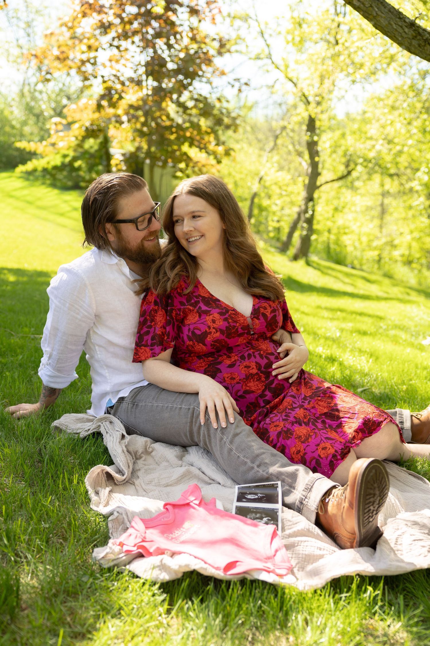 Pregnant couple sitting on a blanket in a sunny park, smiling together with ultrasound photos and pink baby items nearby.