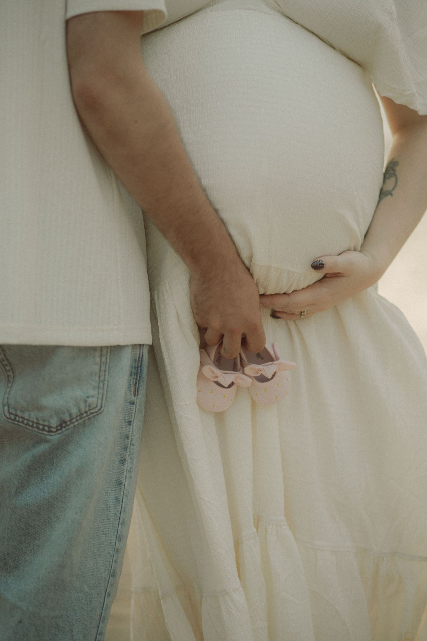 Pregnant couple holding tiny pink baby shoes together, woman in white dress cradling baby bump, maternity photo.