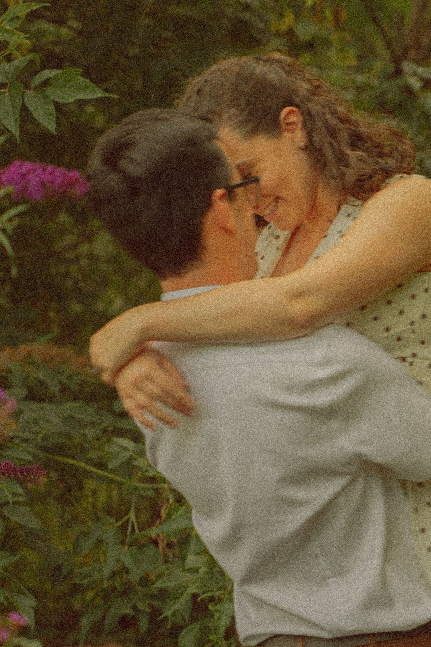 A couple embraces and smiles at each other in a lush garden with pink flowers blooming in the background.