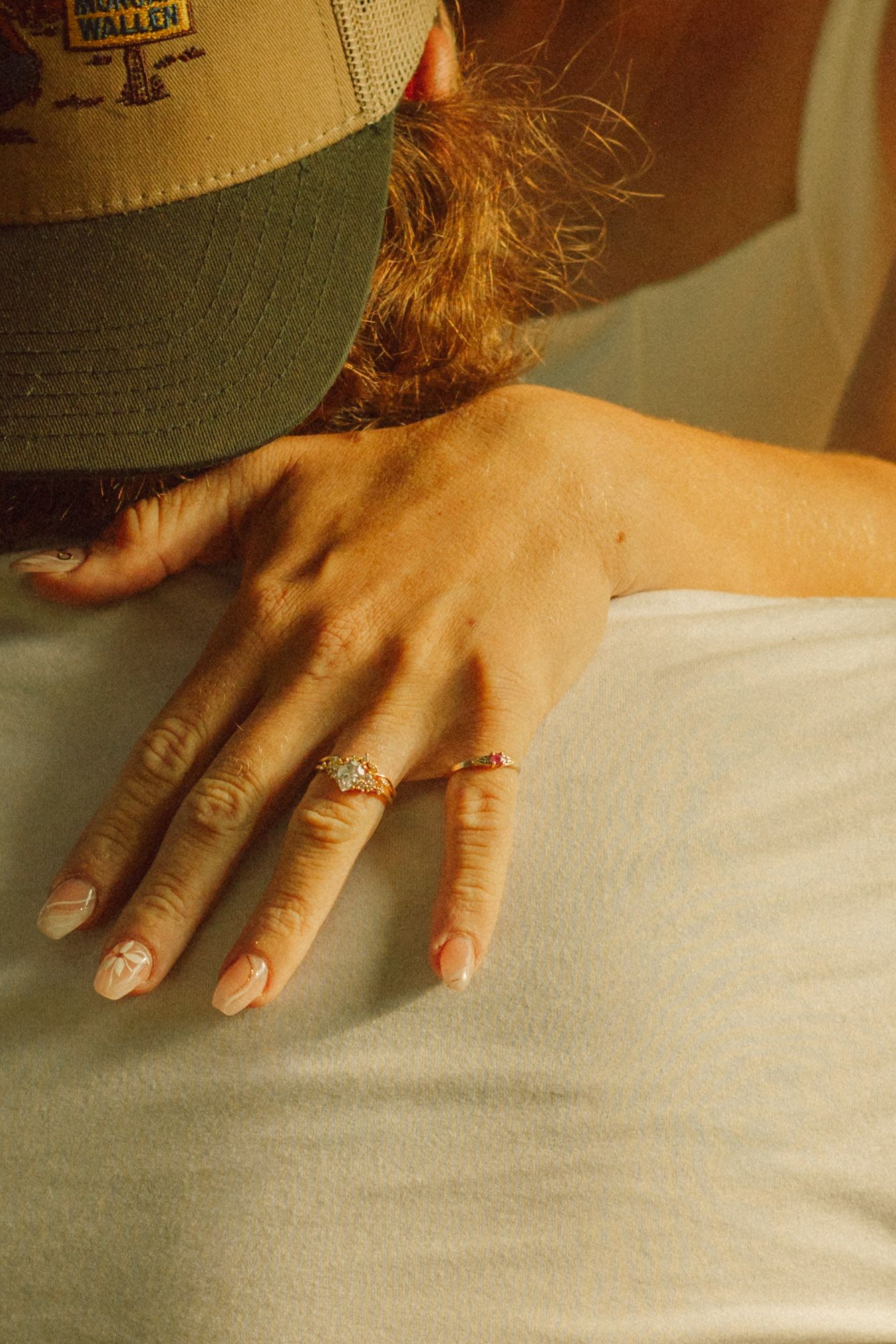 Close-up of a hand wearing a delicate diamond ring, resting on white fabric near a green cap.