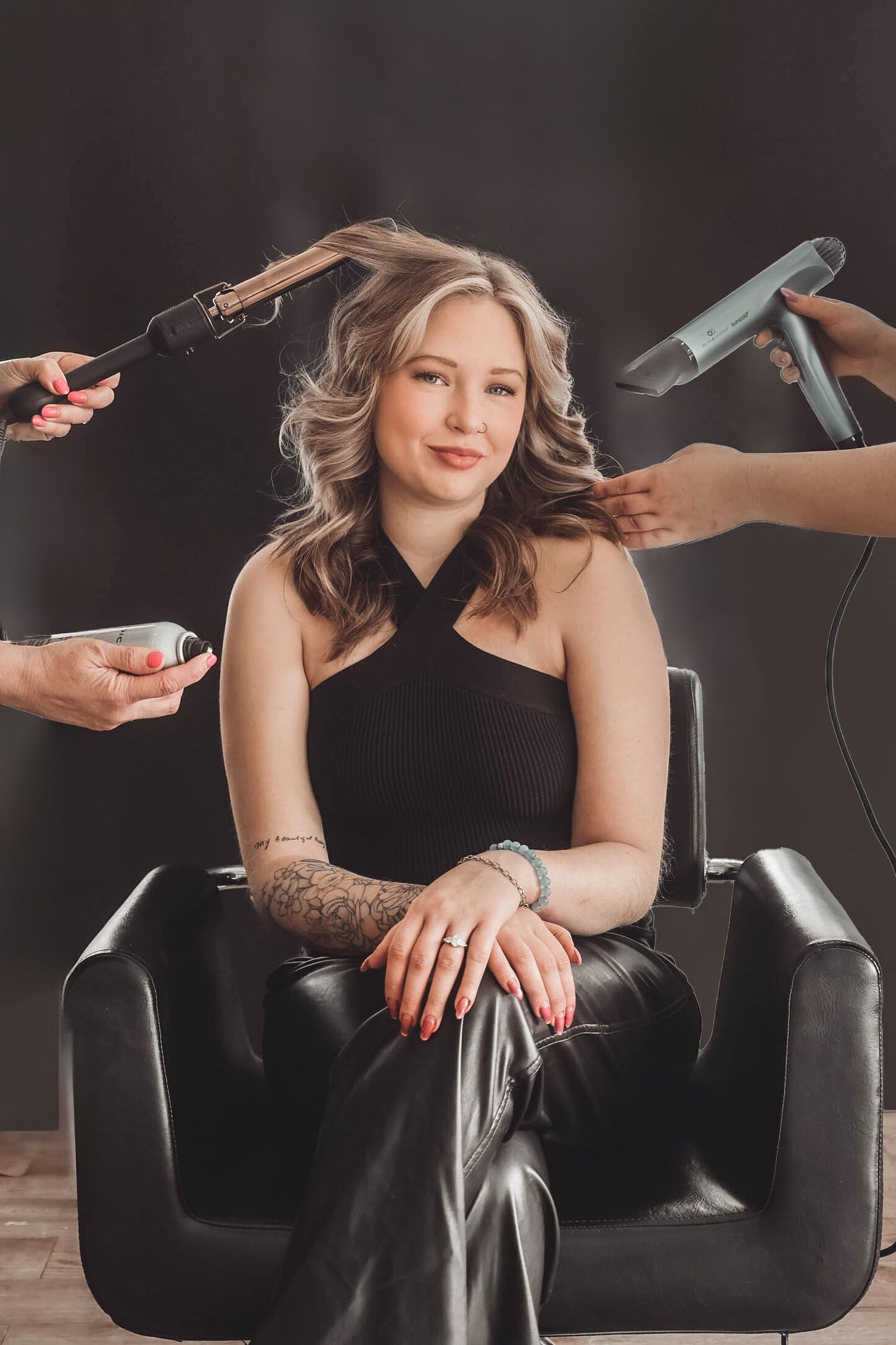 Professional hair styling scene with hands holding various styling tools around a seated figure in black.