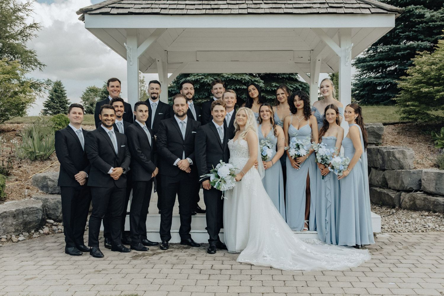 Wedding party poses under white gazebo, bride in white gown with groomsmen in black suits and bridesmaids in blue dresses.