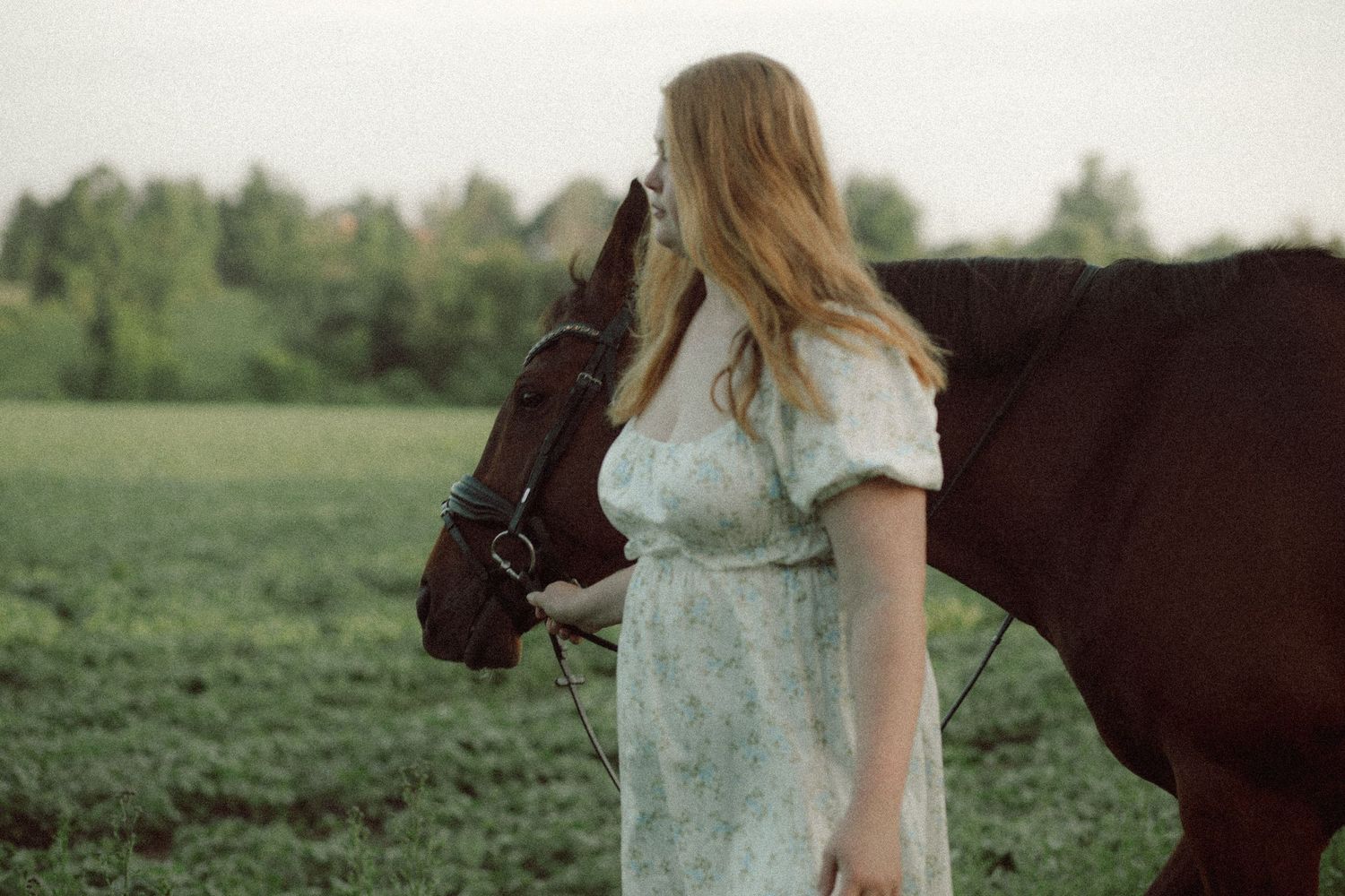 A young woman in a floral dress stands in a green field, gently holding the reins of a brown horse.