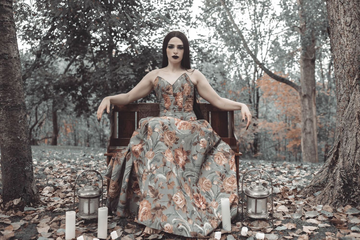 A woman in a floral dress poses gracefully in a rustic outdoor setting among trees and stone walls.