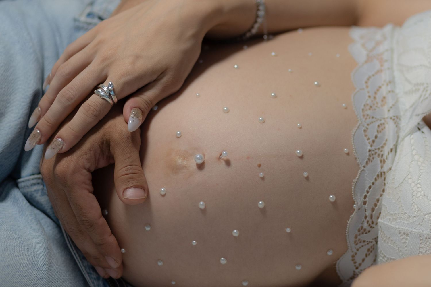 Close-up of pregnant belly decorated with pearl studs, hands gently resting on bump.