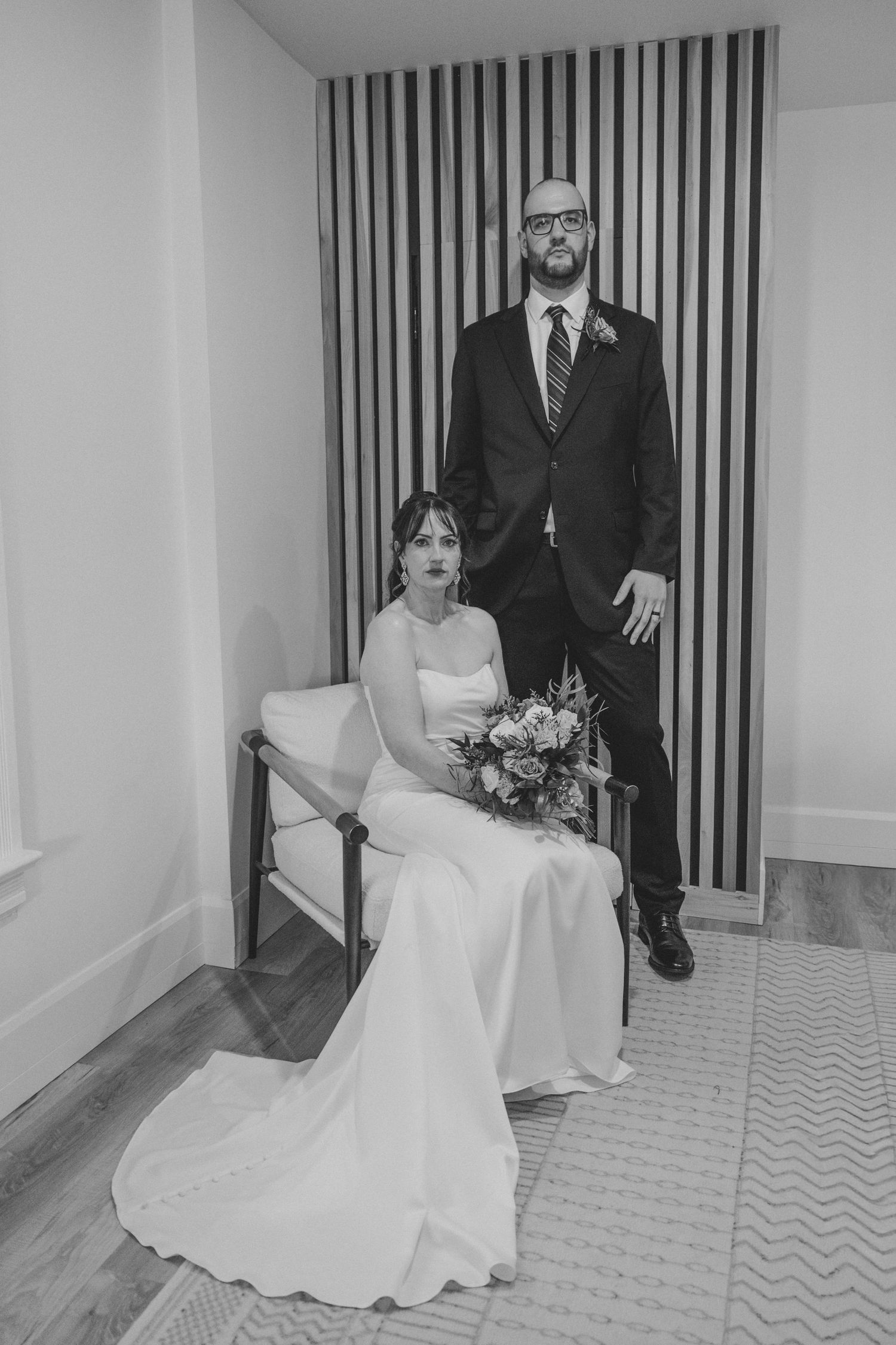 Black and white portrait of bride seated with bouquet and groom standing beside her.