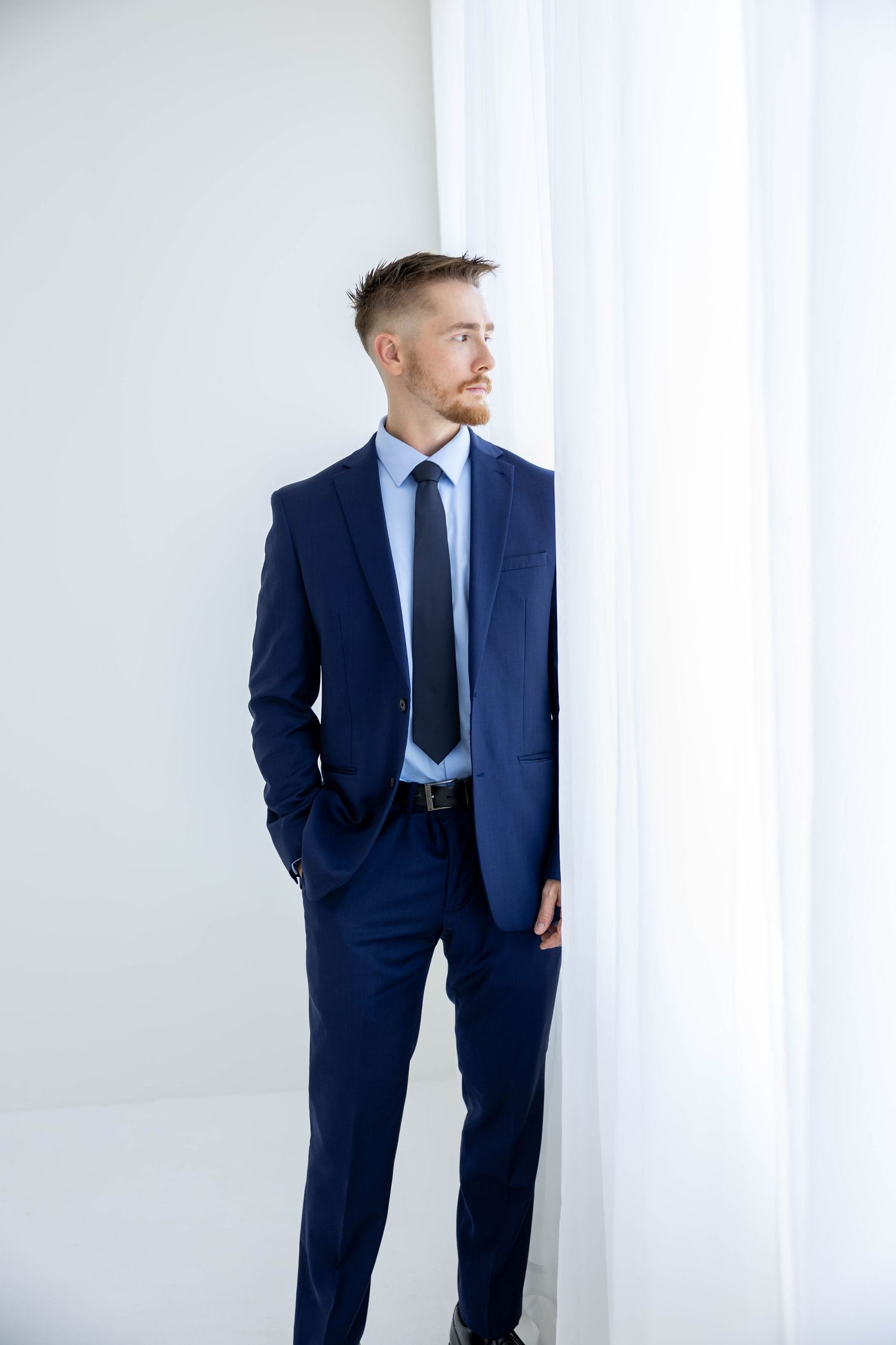 Professional wearing a navy blue suit and tie stands against white curtain background in business setting.