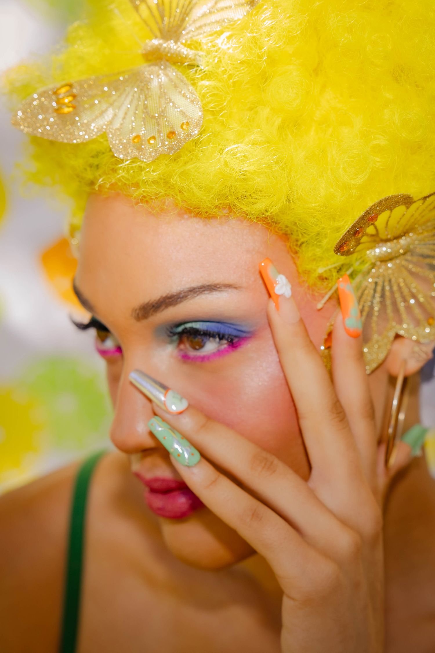 Artistic beauty portrait with bright yellow hair, colorful eye makeup, and pastel blue nails against blurred background.