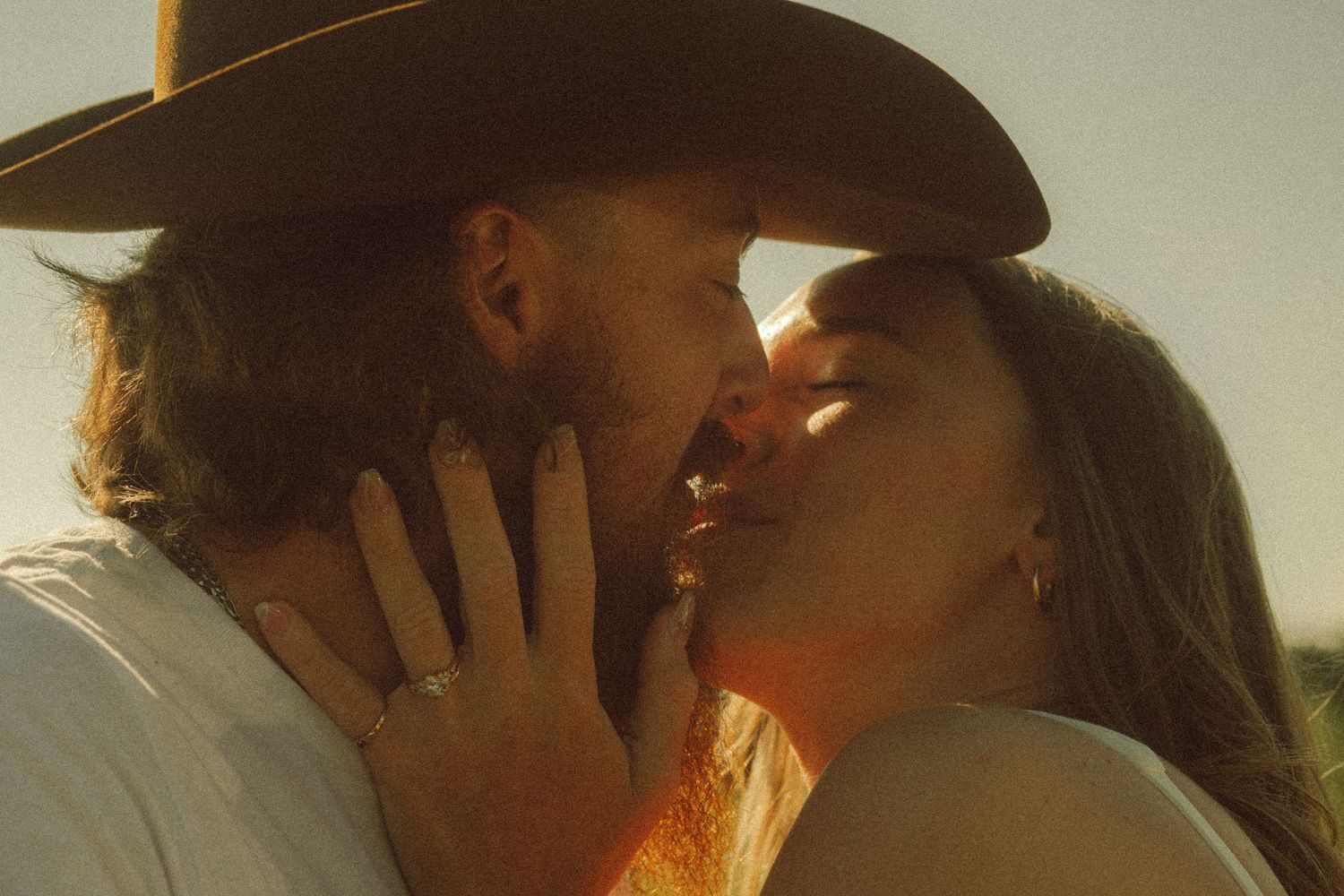 Couple sharing a romantic kiss at golden hour, woman wearing an engagement ring and man in cowboy hat.