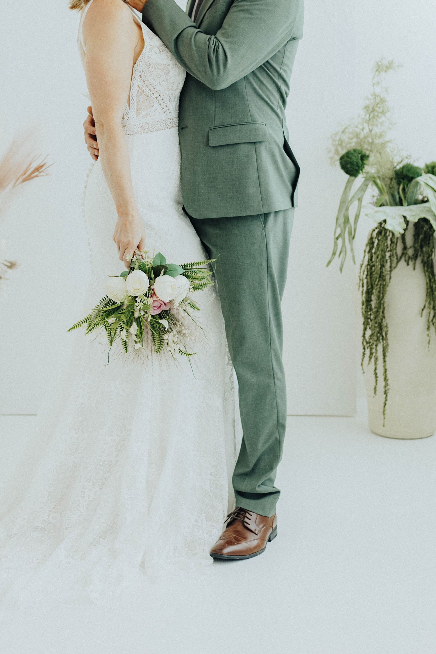 Close-up view of newlyweds with sage green suit and white lace dress holding floral bouquet against white backdrop.