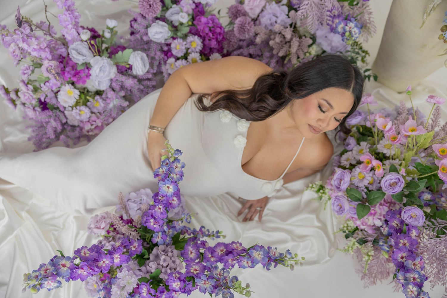 A dreamy portrait of someone lying on a bed surrounded by purple lilac flowers and soft white bedding.