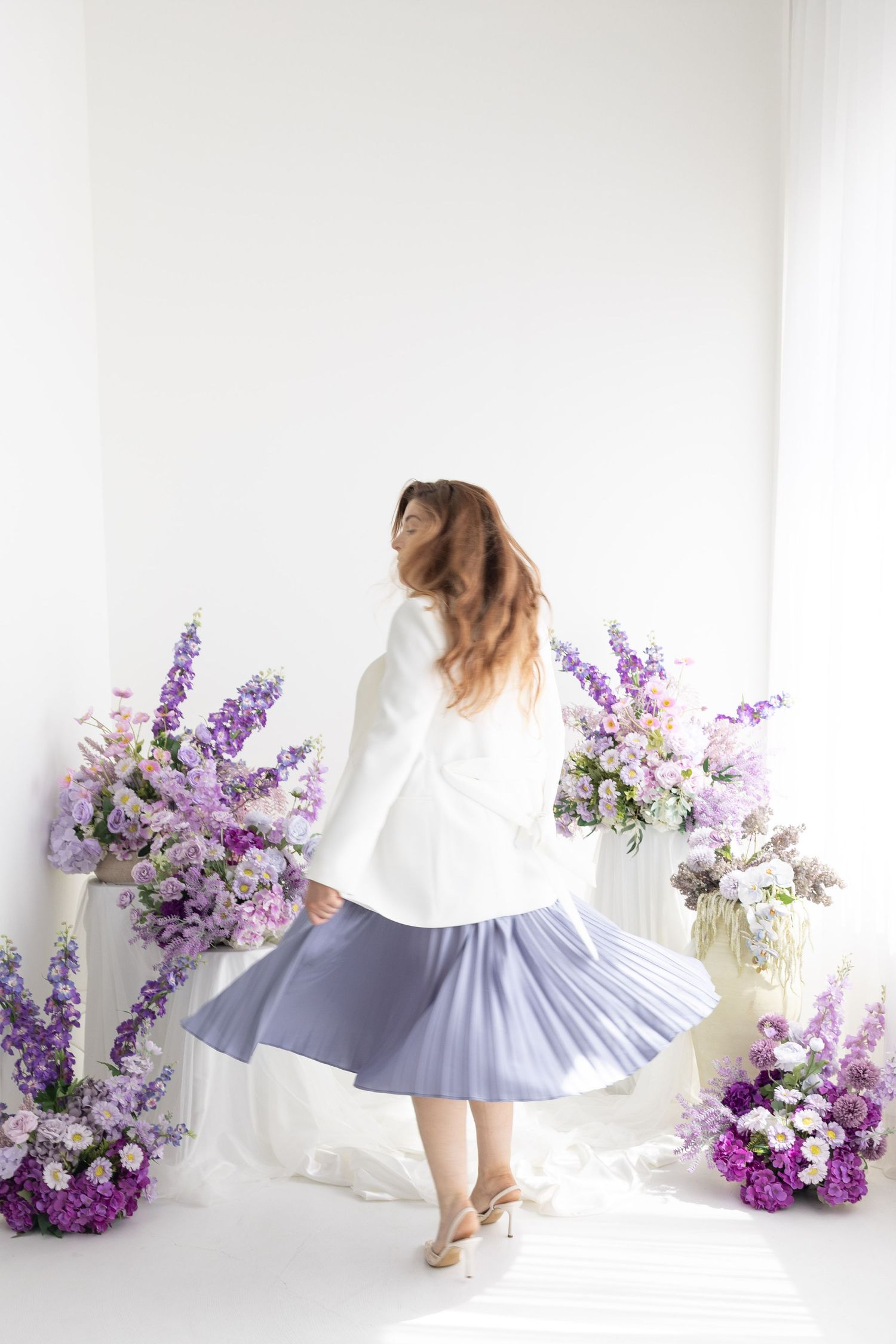 Woman in white blazer and blue pleated skirt twirling among purple floral arrangements in bright white studio.