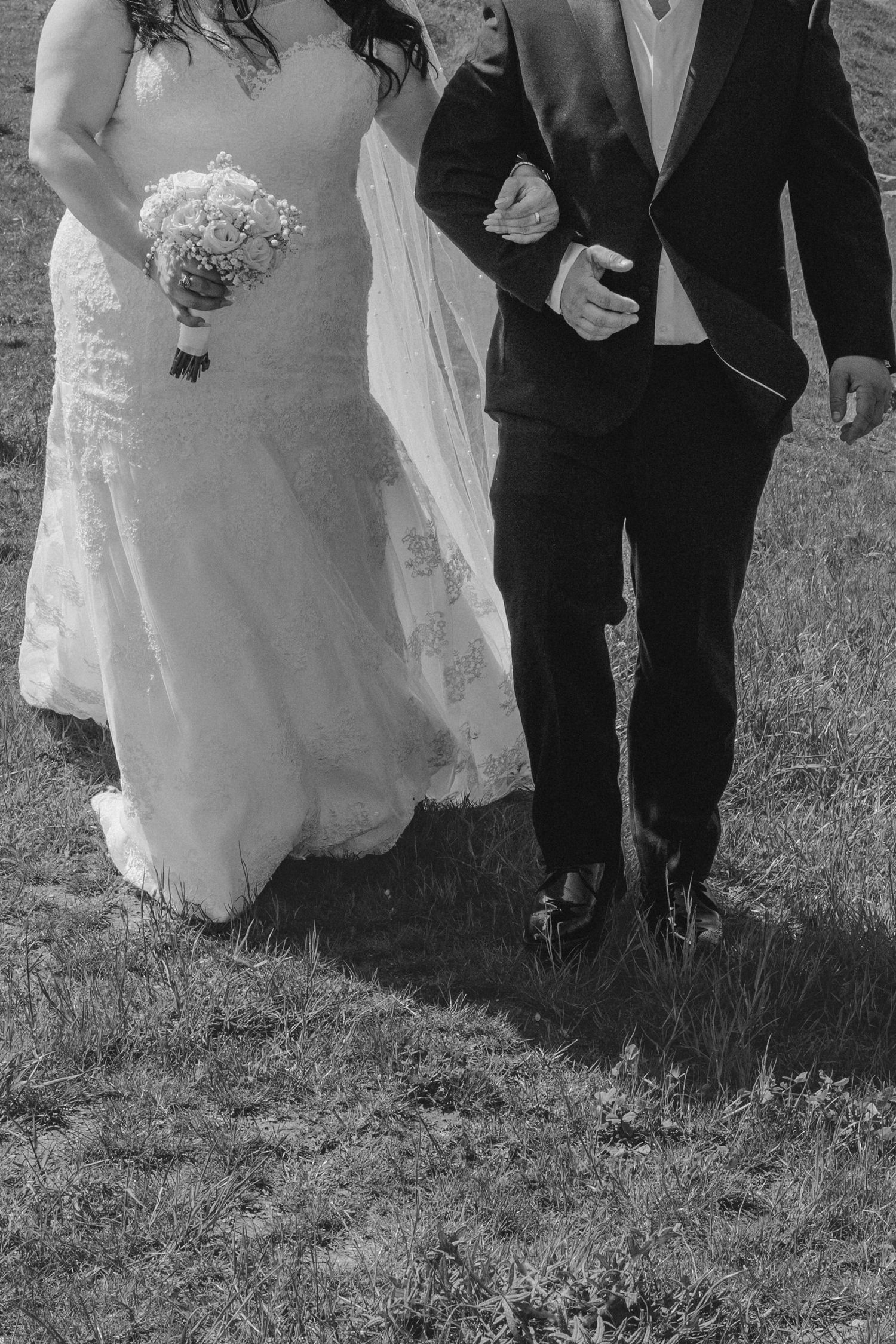 Black and white photo of bride in lace gown holding bouquet walking arm in arm with groom in dark suit on grass.