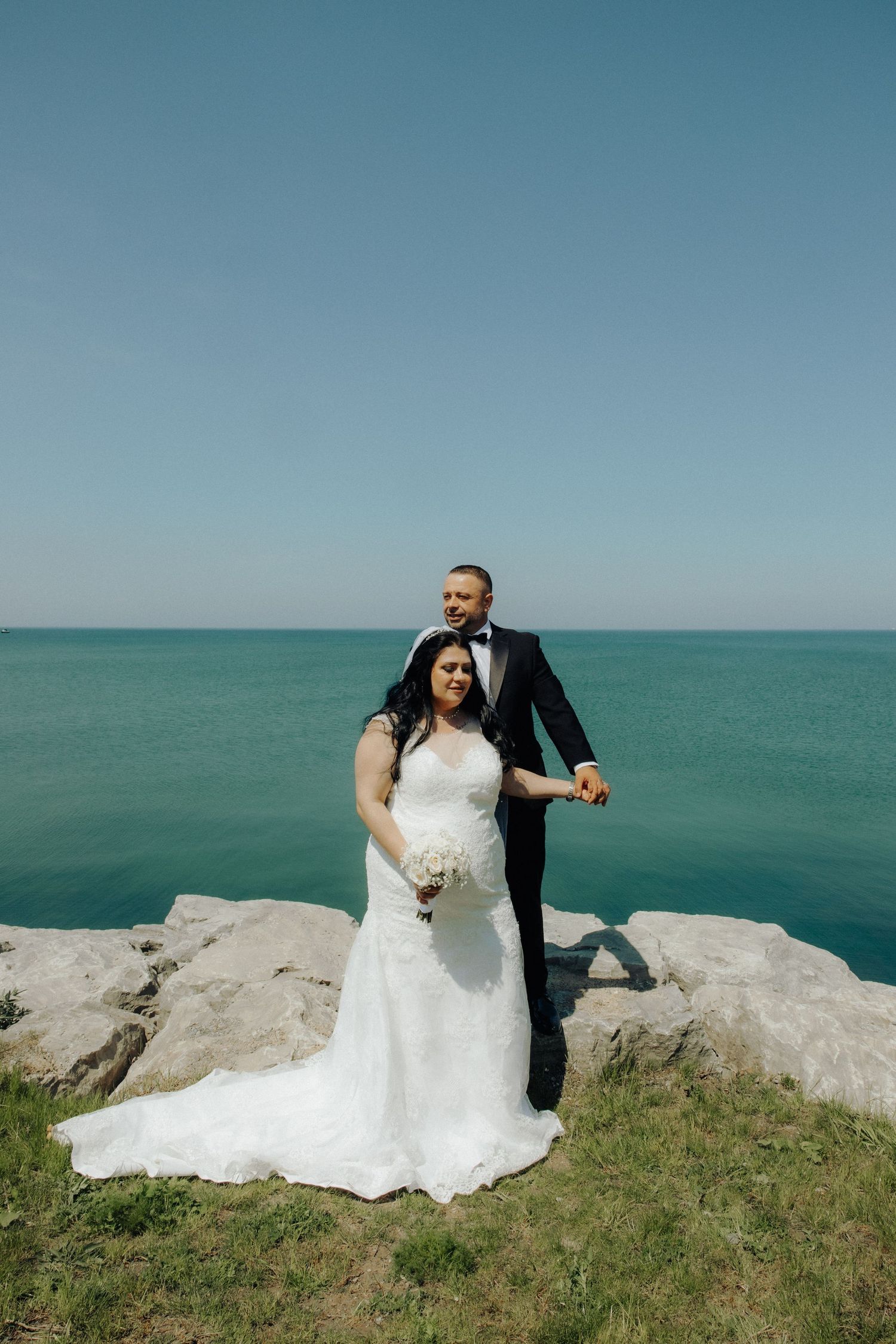 Groom embracing bride holding white bouquet on rocky seaside cliff with turquoise water backdrop.