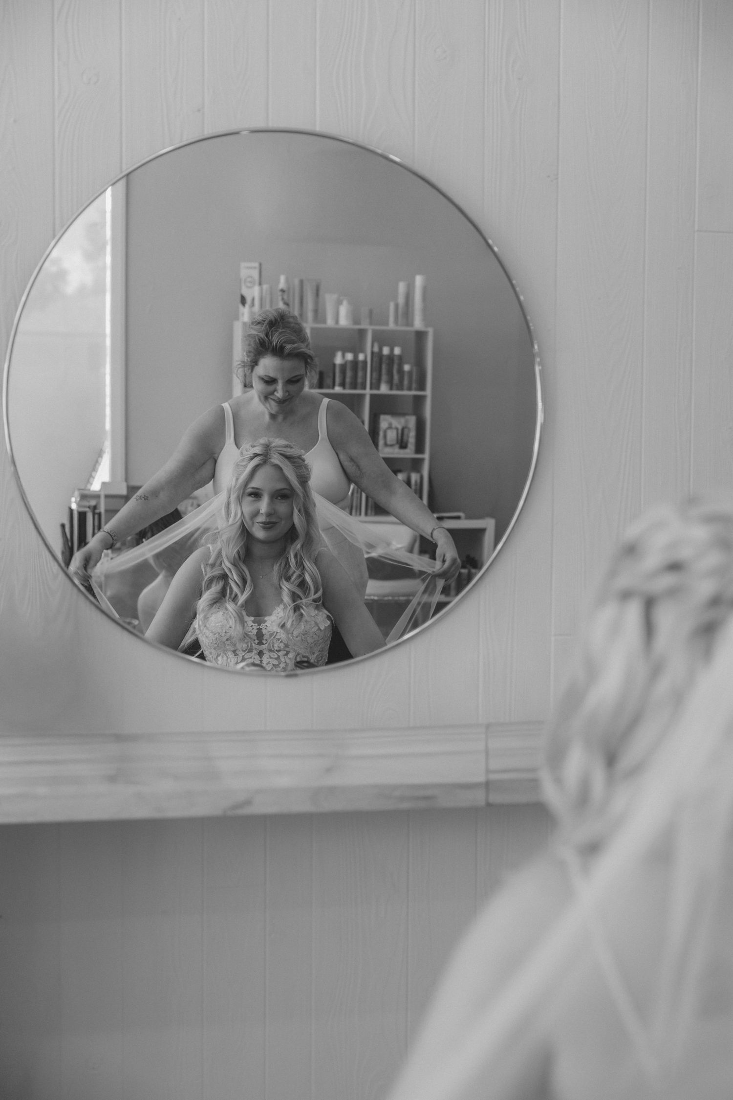 Black and white photo of a bride getting her hair done, reflected in a round salon mirror.