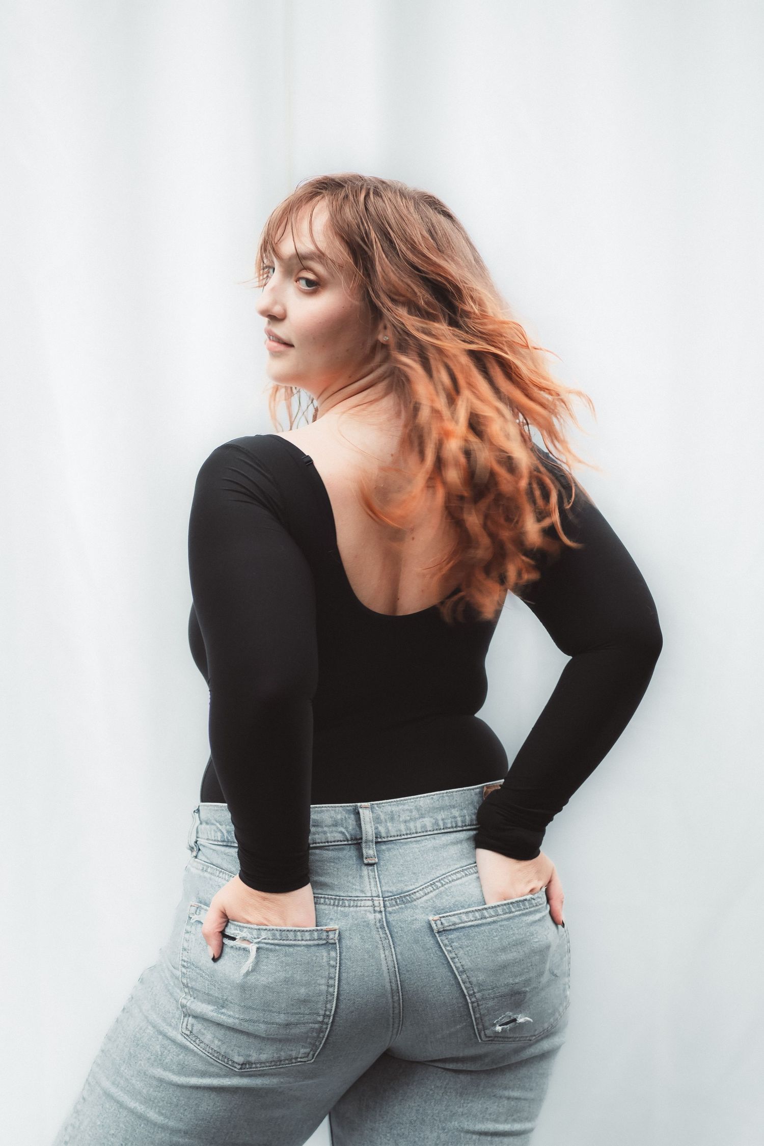 Young woman with wavy auburn hair poses in black backless top and light wash jeans against white background.