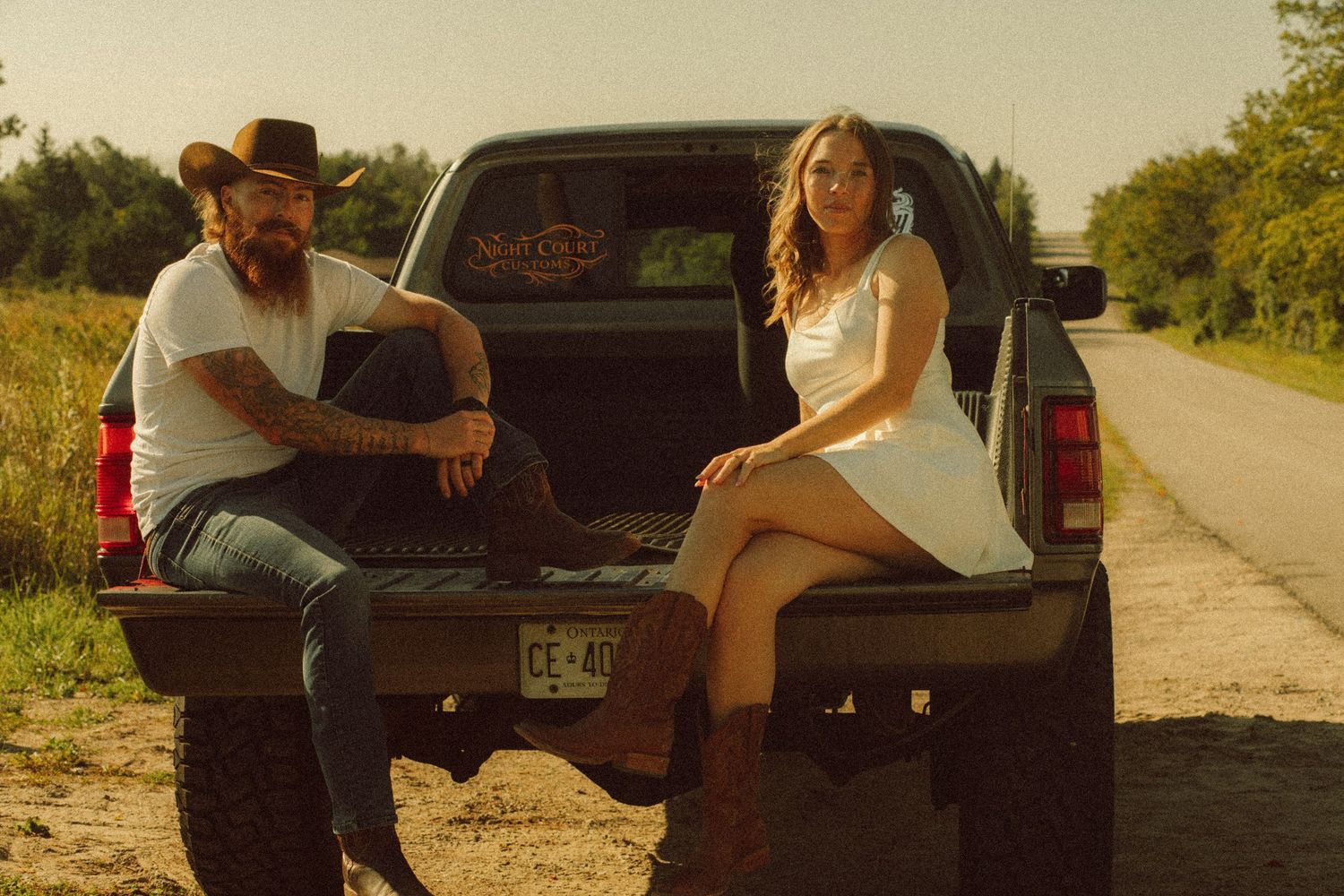 A man and woman sit on a truck tailgate on a rural road at sunset, dressed in casual country style attire.