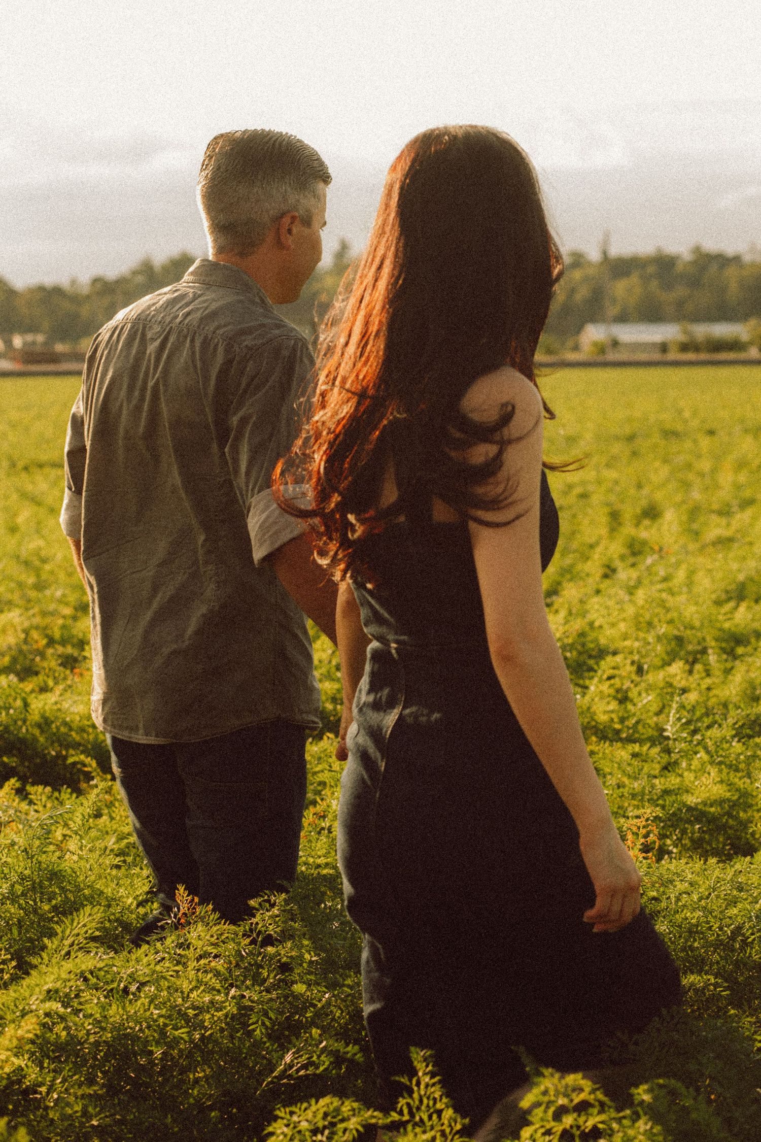 A couple walks hand in hand through a lush green field at golden hour, woman with red hair in dark dress.