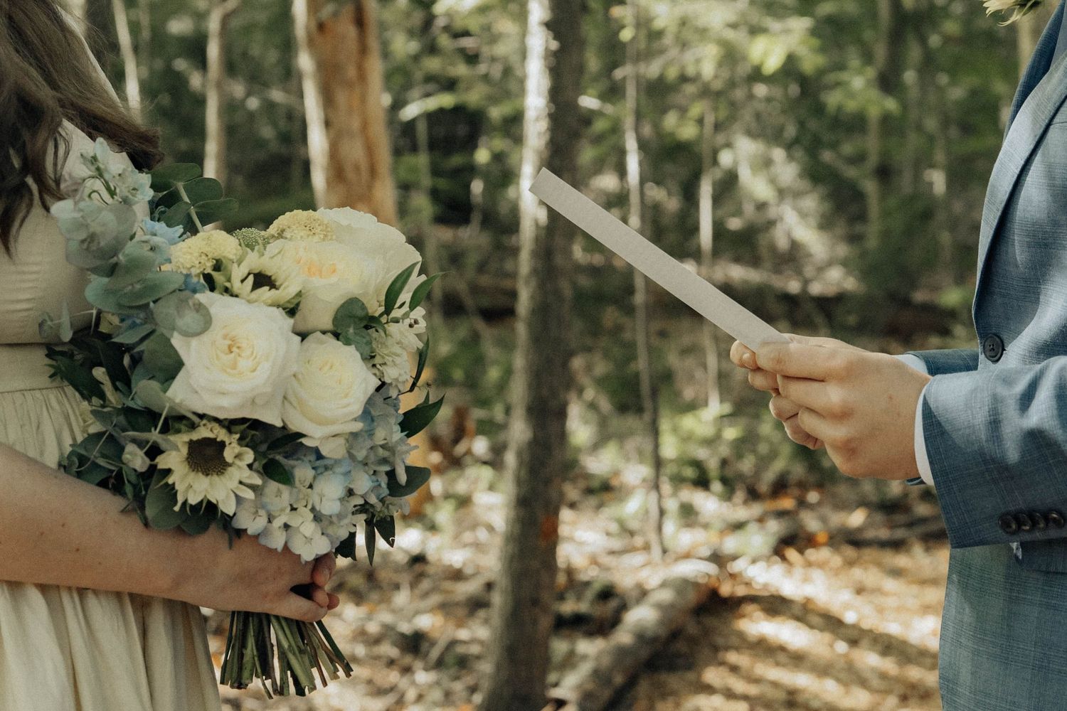 A couple in wedding attire holds hands in a serene forest setting surrounded by towering pine trees.
