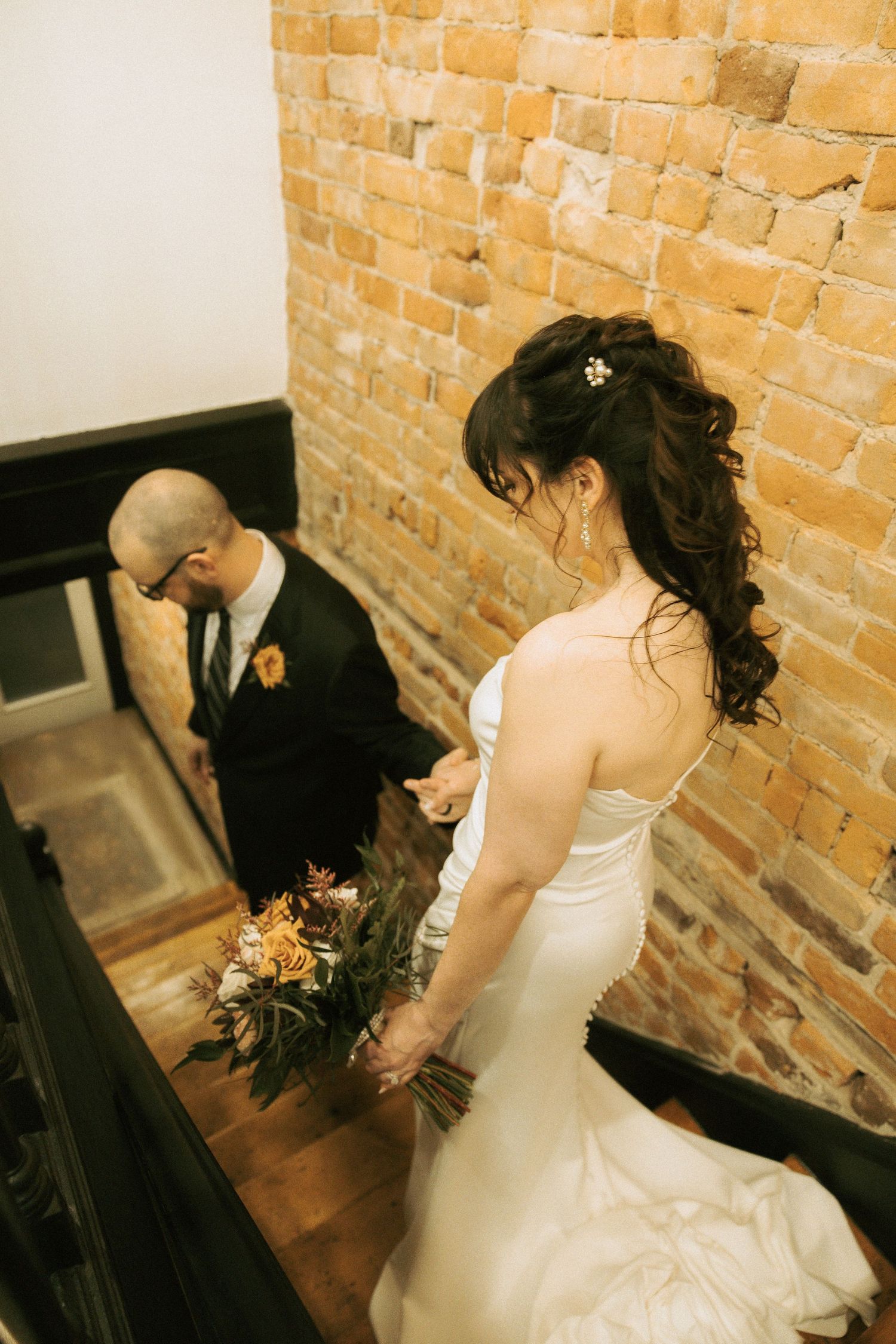 Bride in fitted white gown holds bouquet descending wooden staircase with groom near exposed brick wall.