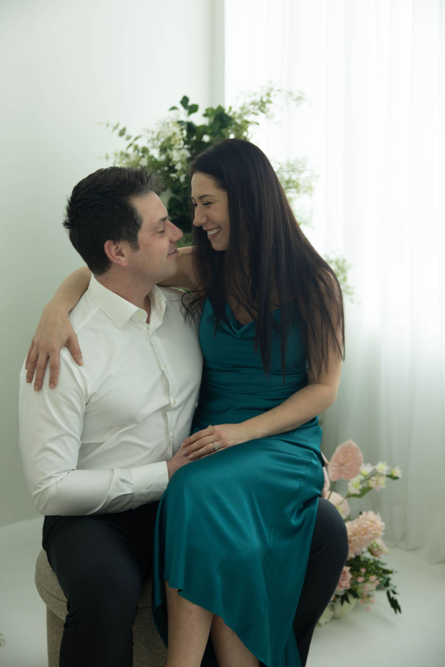 A couple in a white shirt and teal dress share intimate moments in a bright studio setting with floral decor.