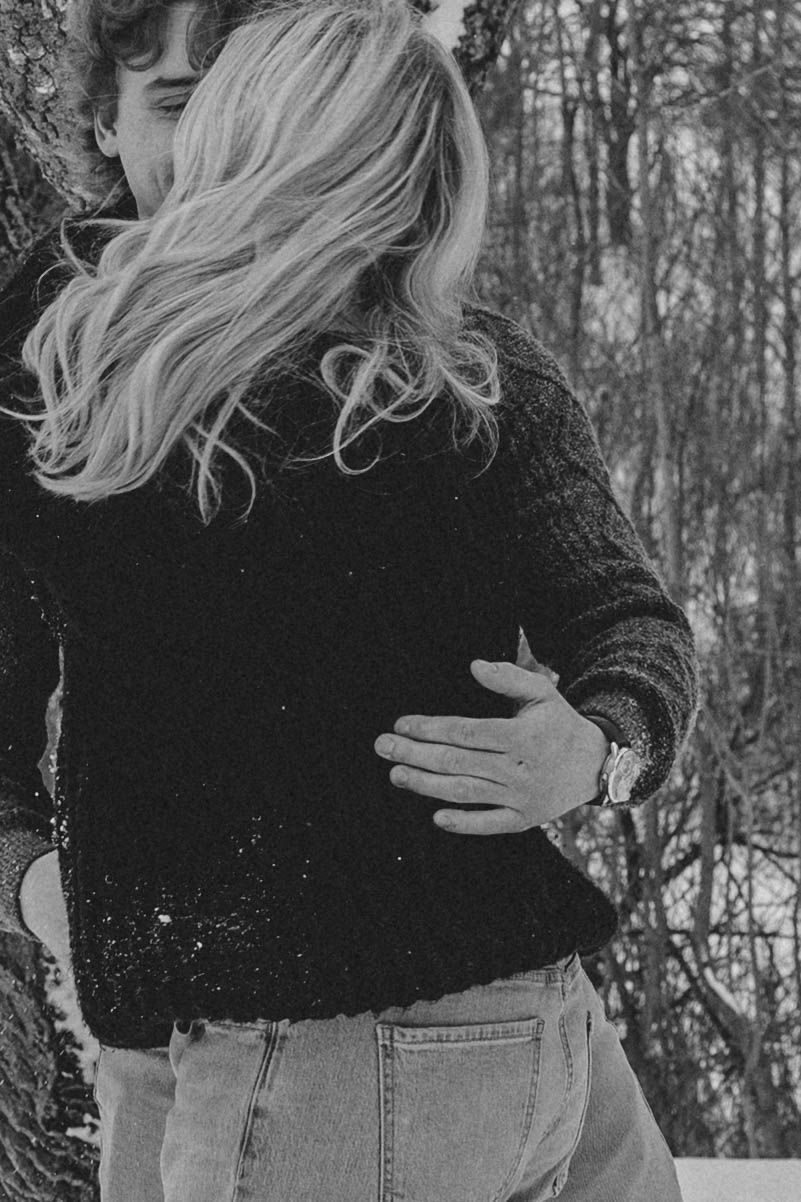 Black and white photo showing blonde windswept hair and casual winter outfit with jeans against snowy forest backdrop.