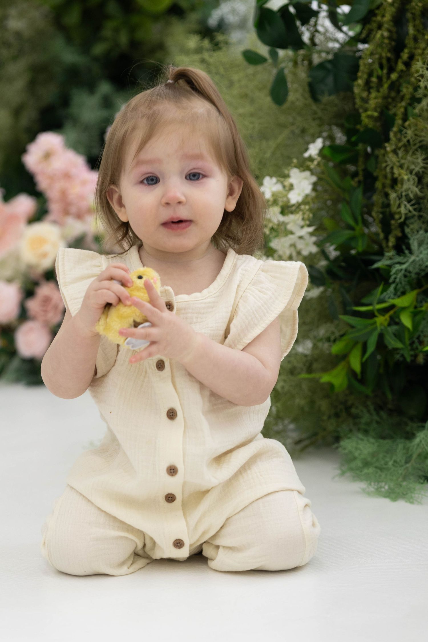 A toddler in a cream-colored button-up romper sits on a white backdrop with pink roses and greenery in the background.