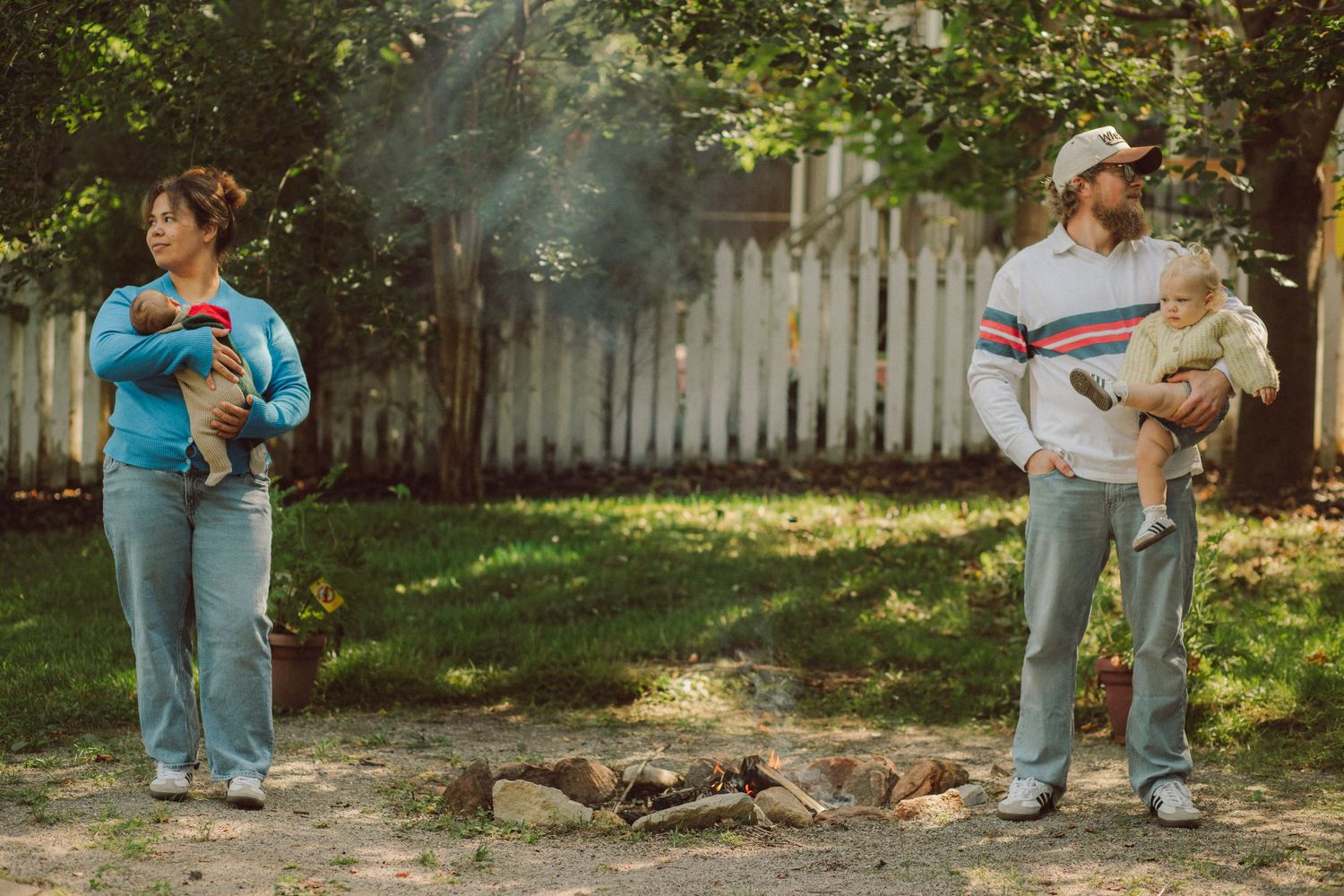 A man and woman stand apart in a backyard near a campfire, each holding a small animal, with a white fence behind them.