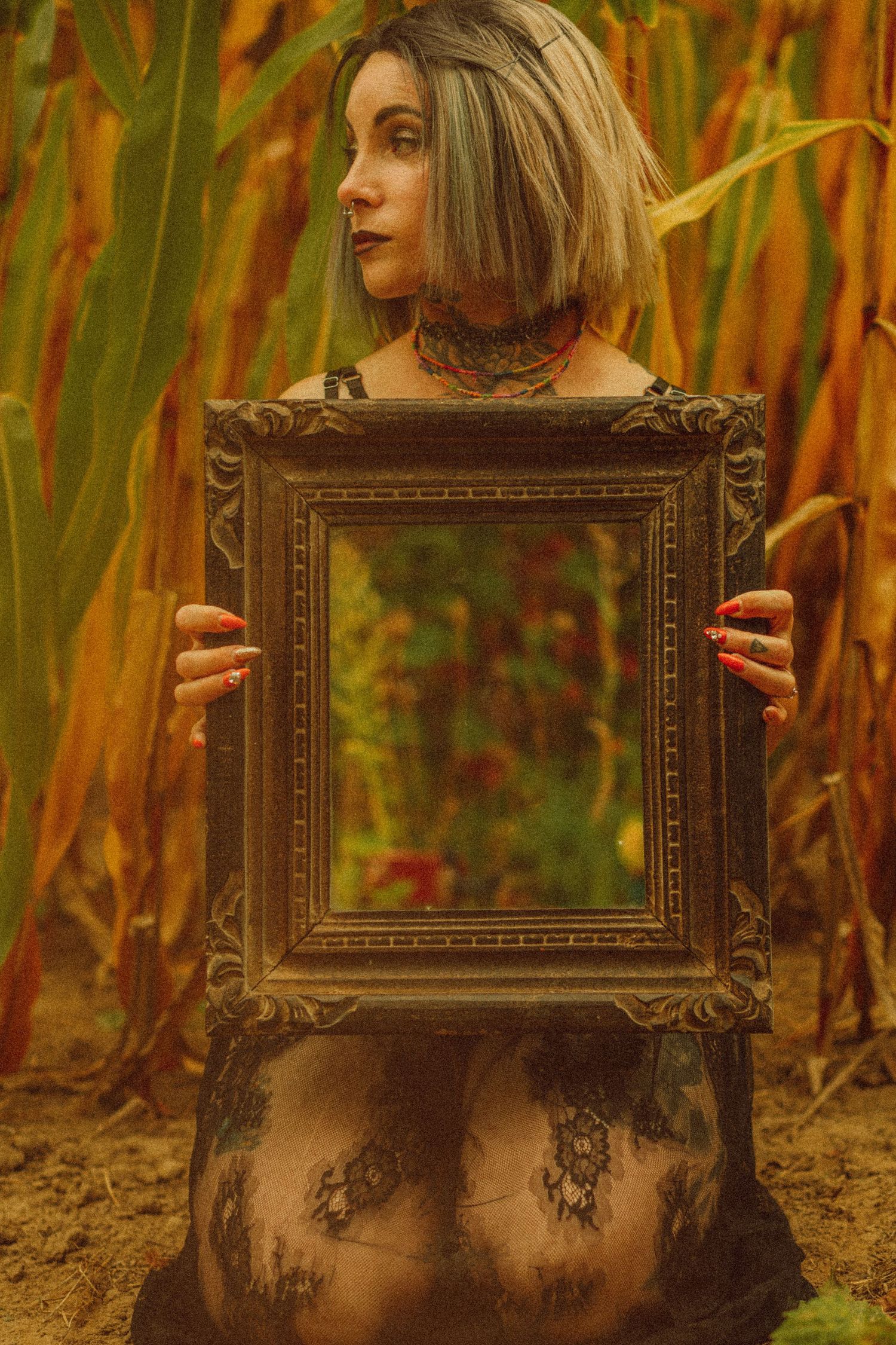 Tattooed woman holding ornate vintage mirror frame in a golden cornfield, wearing lace outfit with red nails.