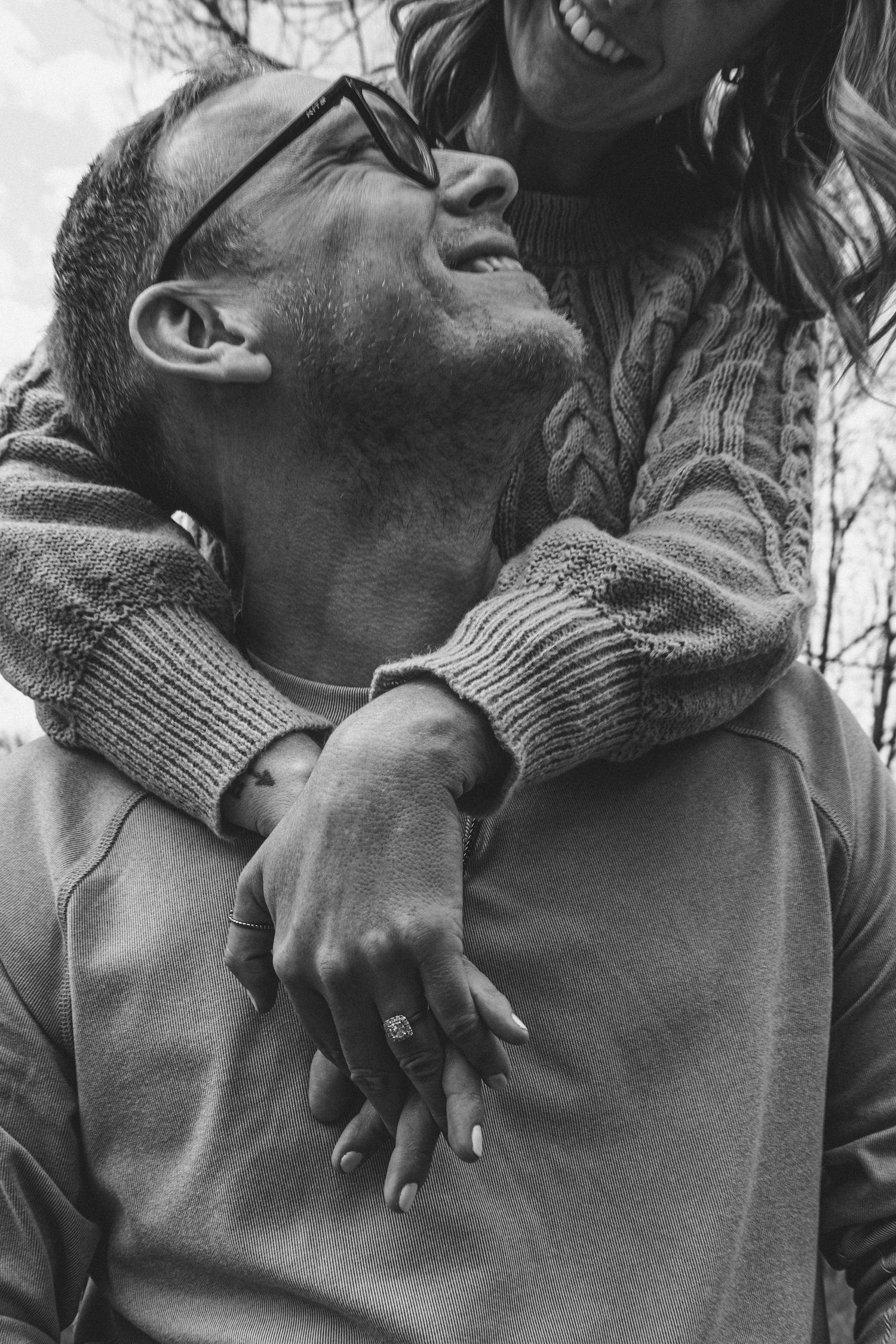 Black and white photo of a happy couple embracing outdoors, woman hugging man from behind showing engagement ring.