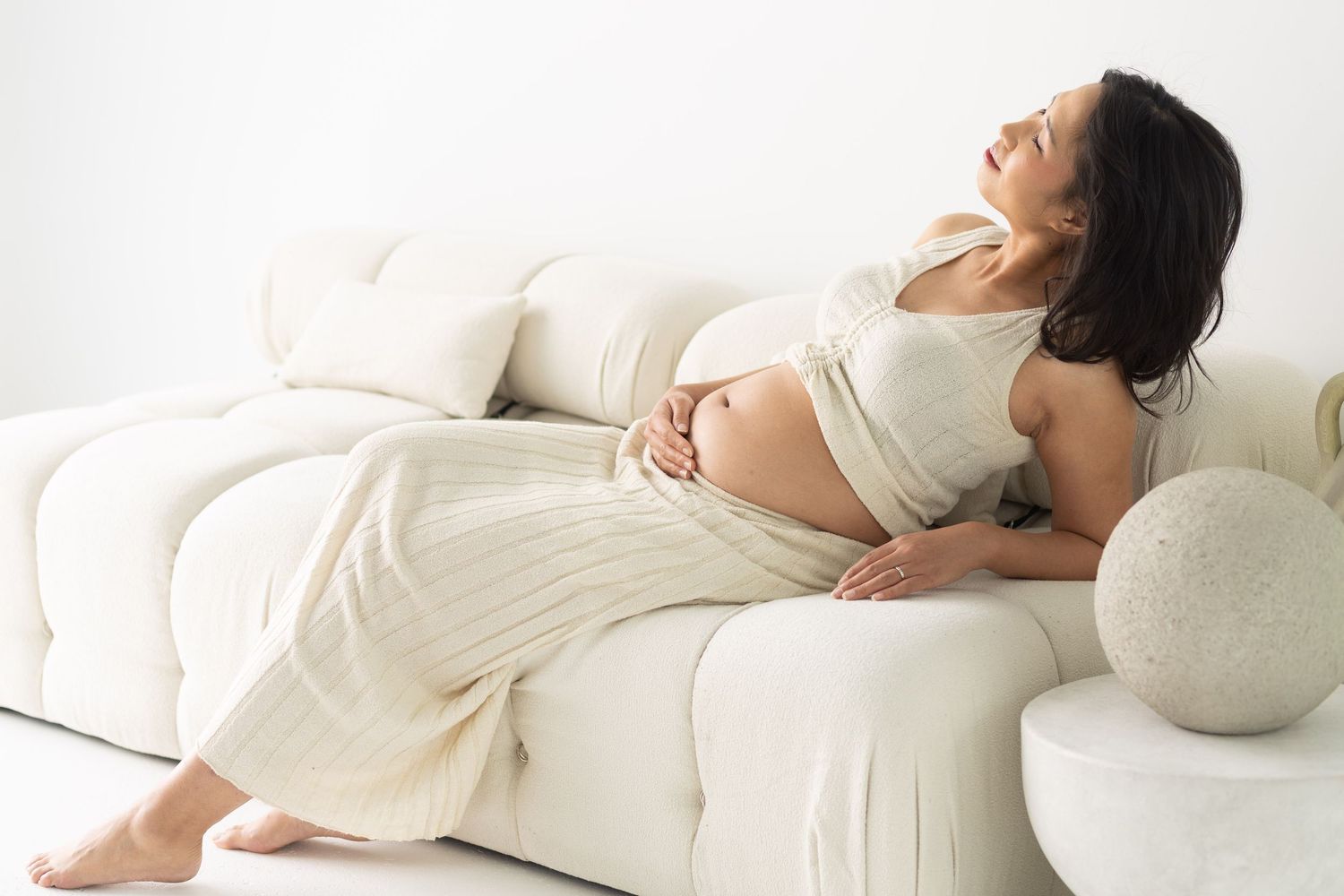 Pregnant woman in cream outfit relaxing on white sofa, cradling baby bump with eyes closed.