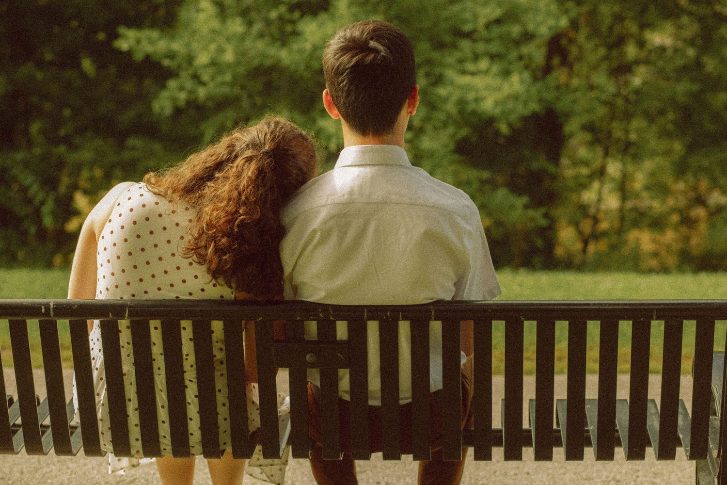 Couple sitting together on a wooden park bench, viewed from behind, surrounded by lush green trees.