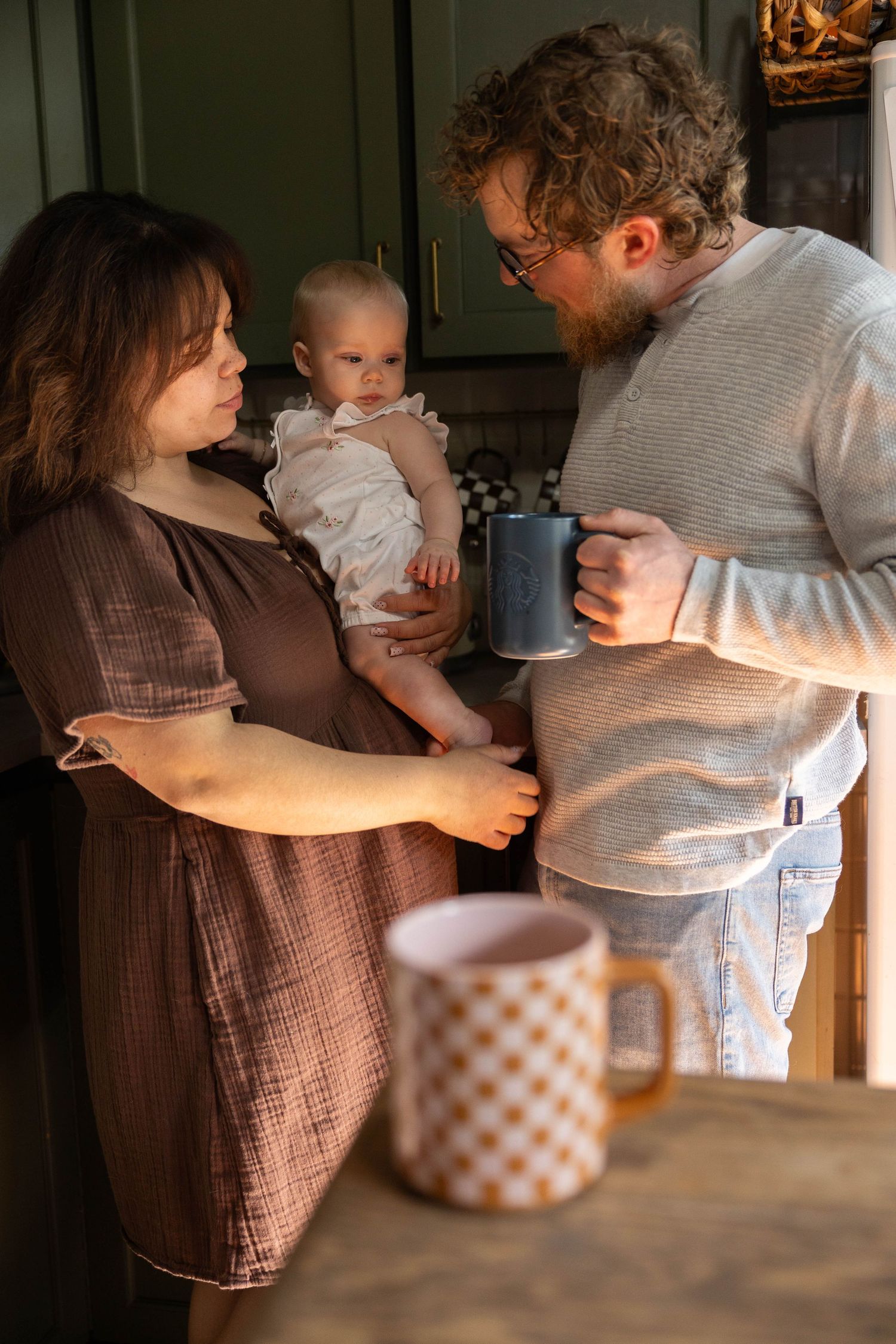 Family moment in kitchen with warm lighting as parents hold child and share a mug together.