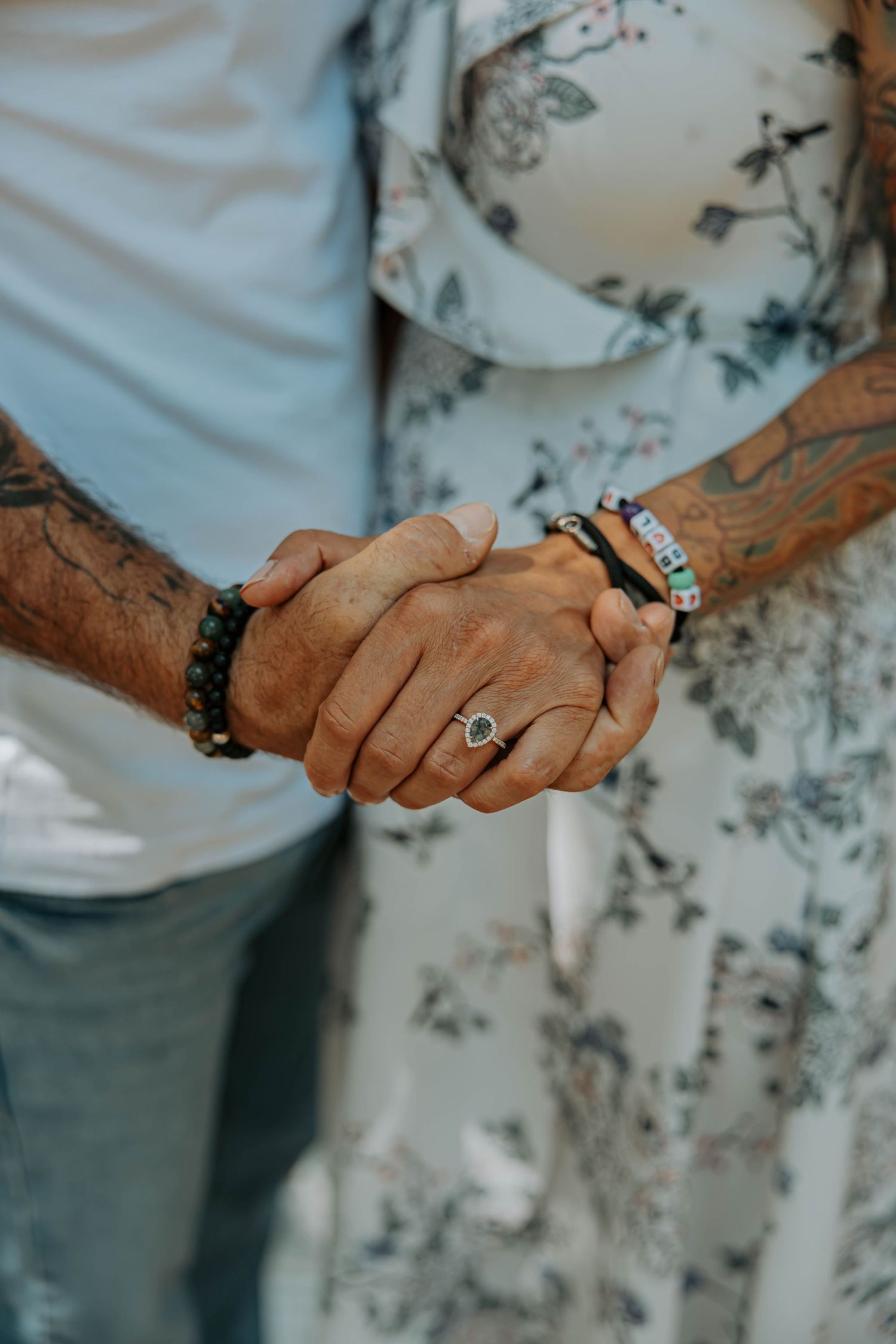 Two hands clasped together showing wedding rings and bracelets against a floral patterned background.