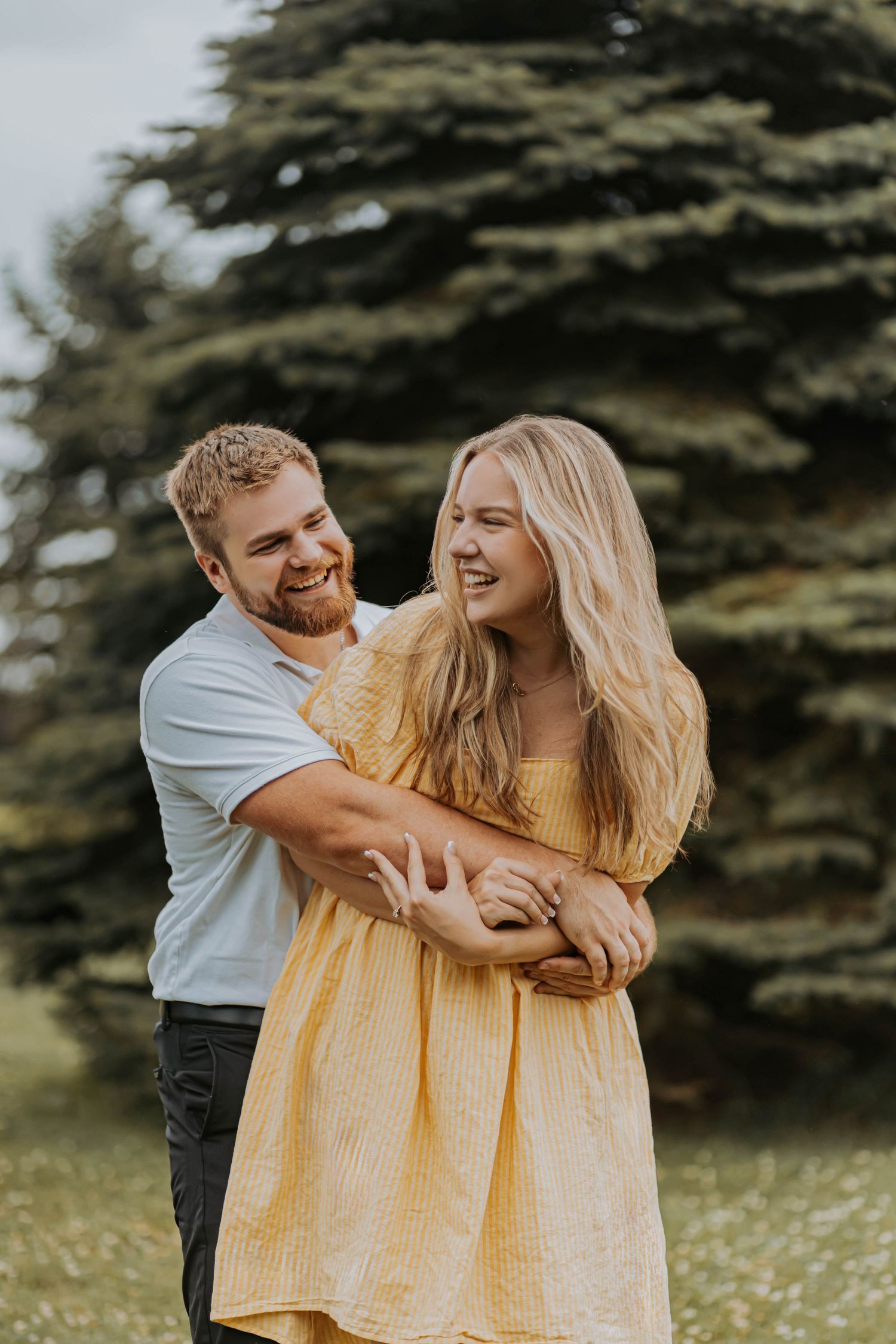Young couple in a field share affectionate moments against evergreen trees with person wearing yellow dress.