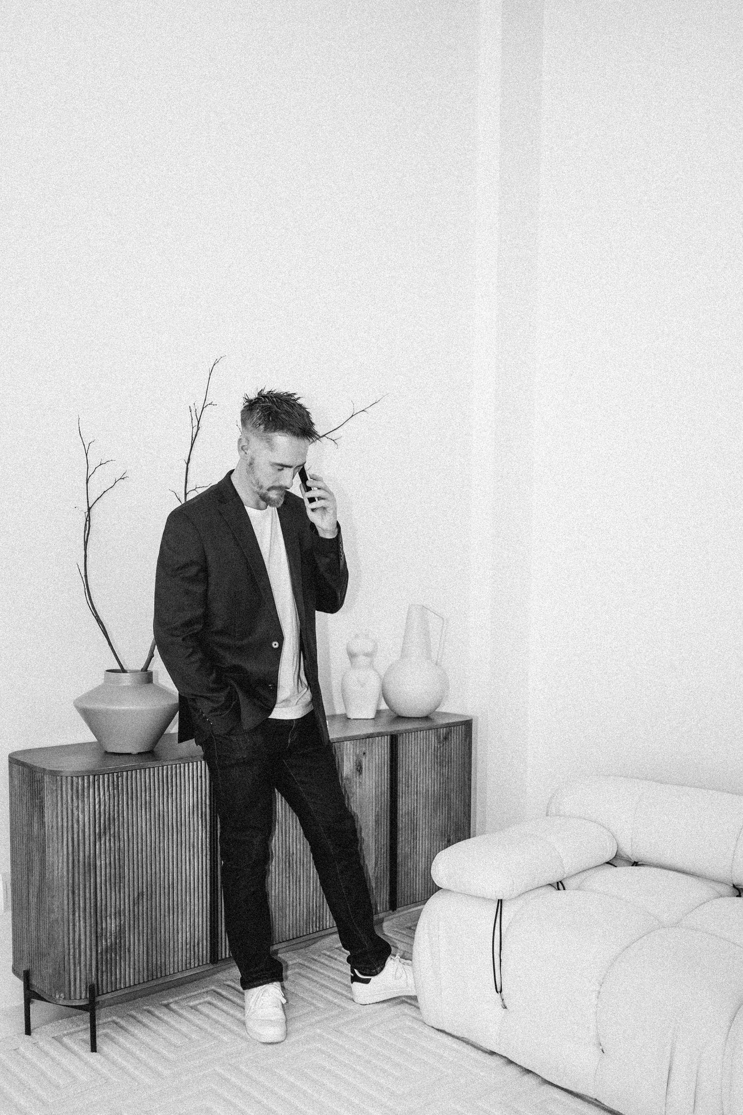 Stylish figure in dark suit poses near modern furniture in minimalist white room setting.