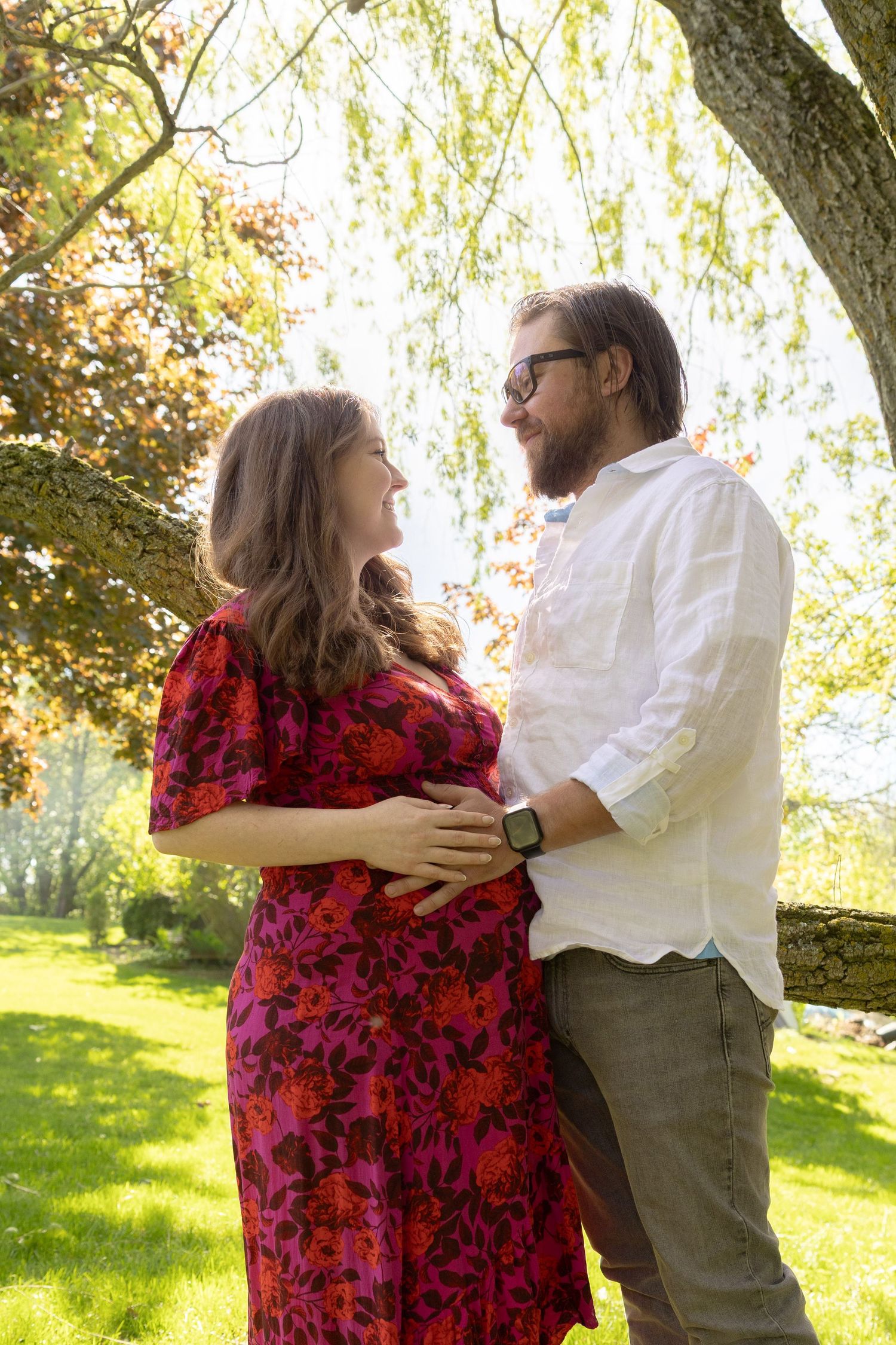 Pregnant couple embracing outdoors under a tree, woman in red floral dress, man in white shirt, smiling at each other.