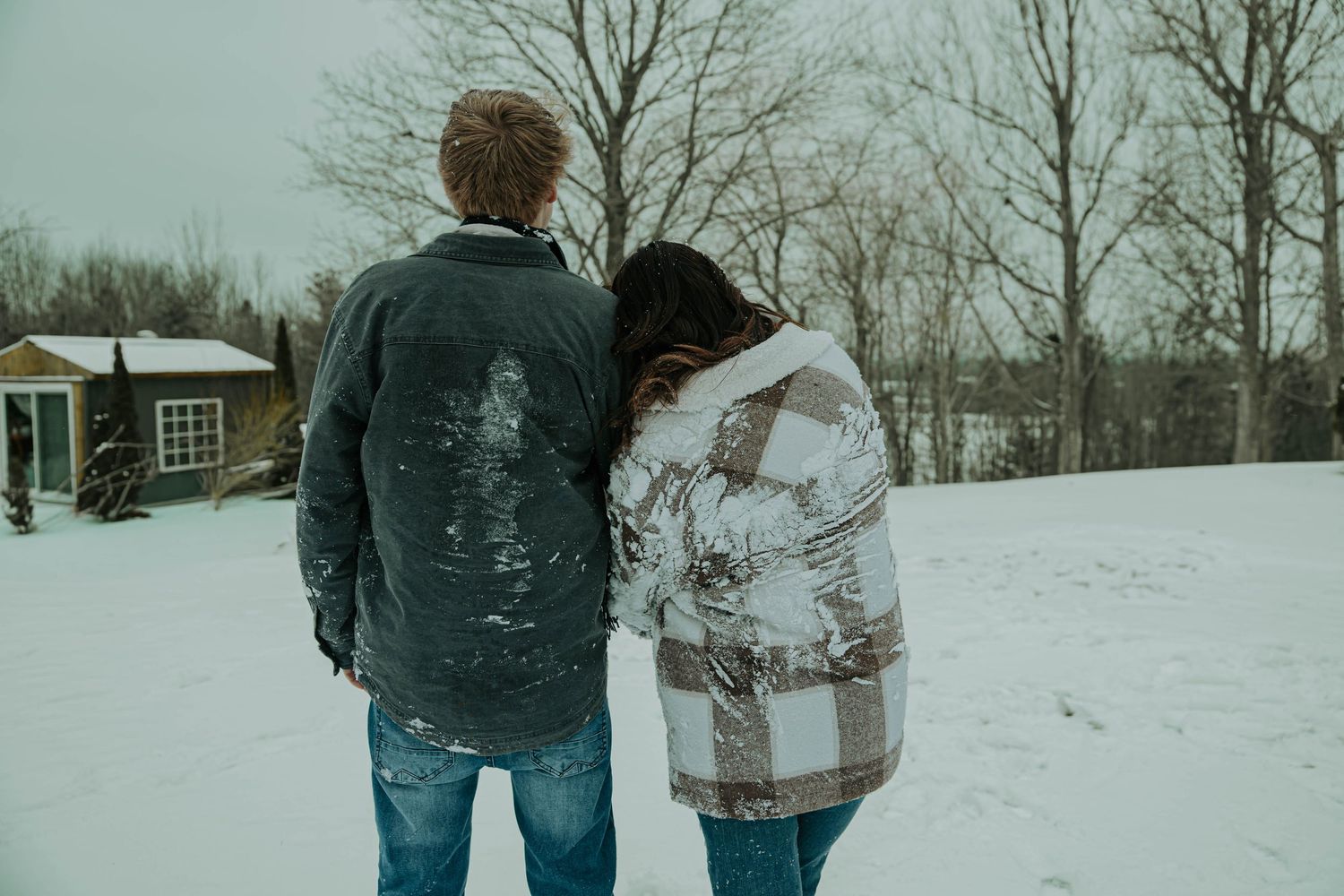 Two people walk together through a snowy winter landscape with bare trees in the background.