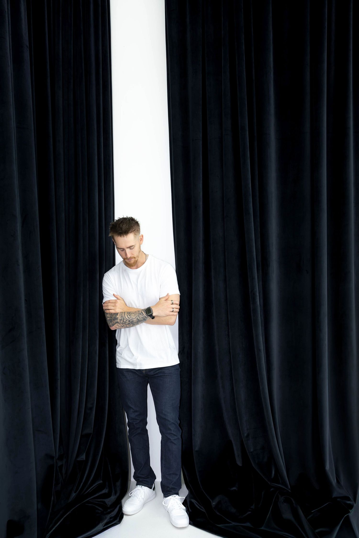 Figure in white t-shirt and dark pants poses against dramatic black curtain backdrop in studio.