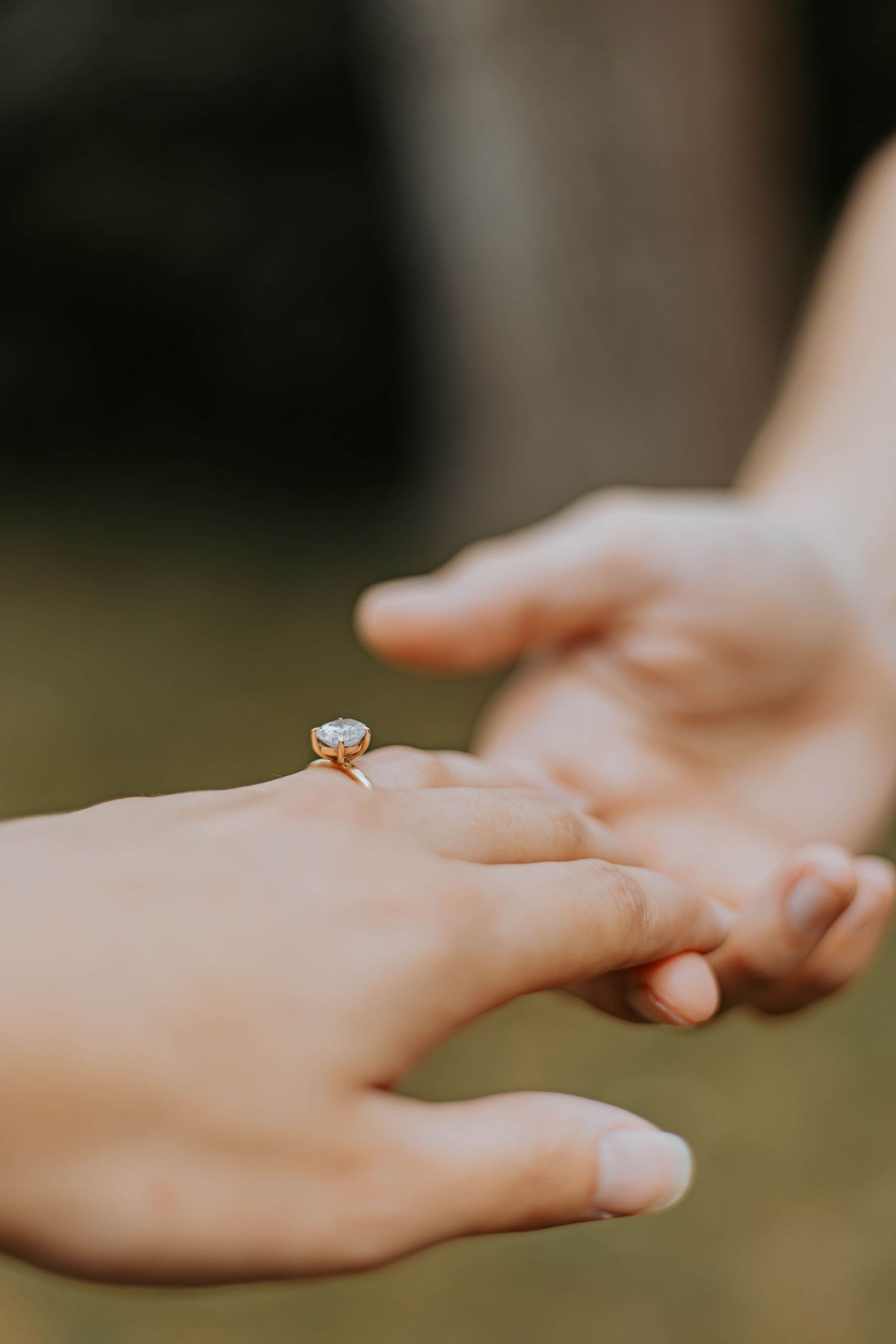Close-up shot of hands wearing a delicate diamond engagement ring against a blurred natural background.