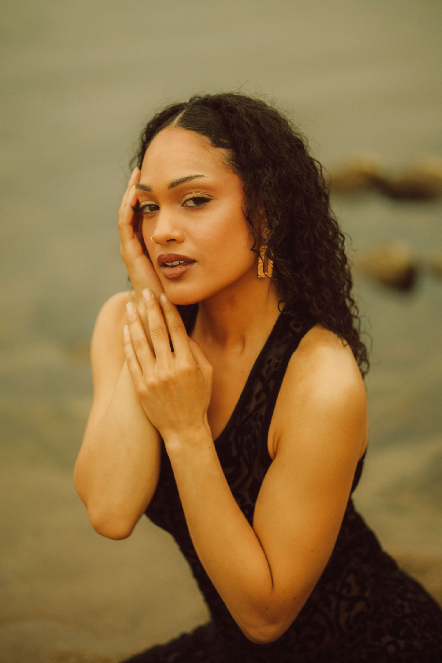 A woman with curly black hair in a black dress poses gracefully by the water, hands near her face.