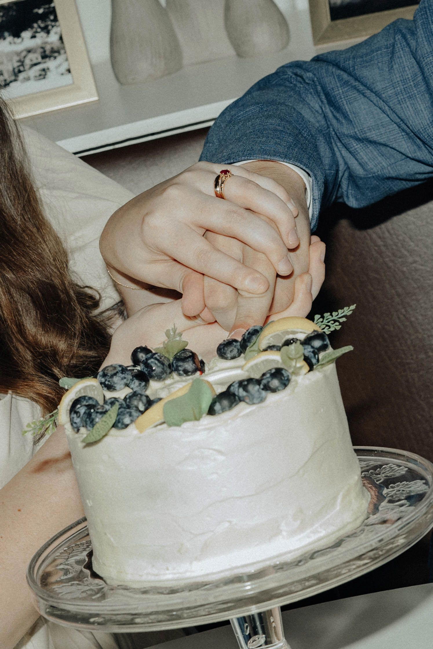 Hands carefully decorating the top of a white frosted cake with dark garnishes and greenery.