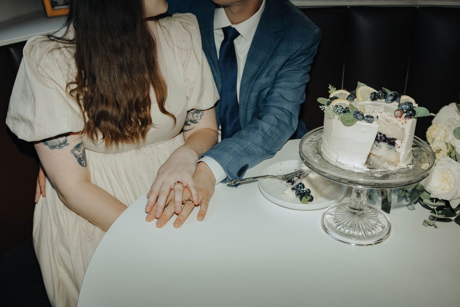 Two hands touch tenderly across a white wedding cake at a reception table setting.